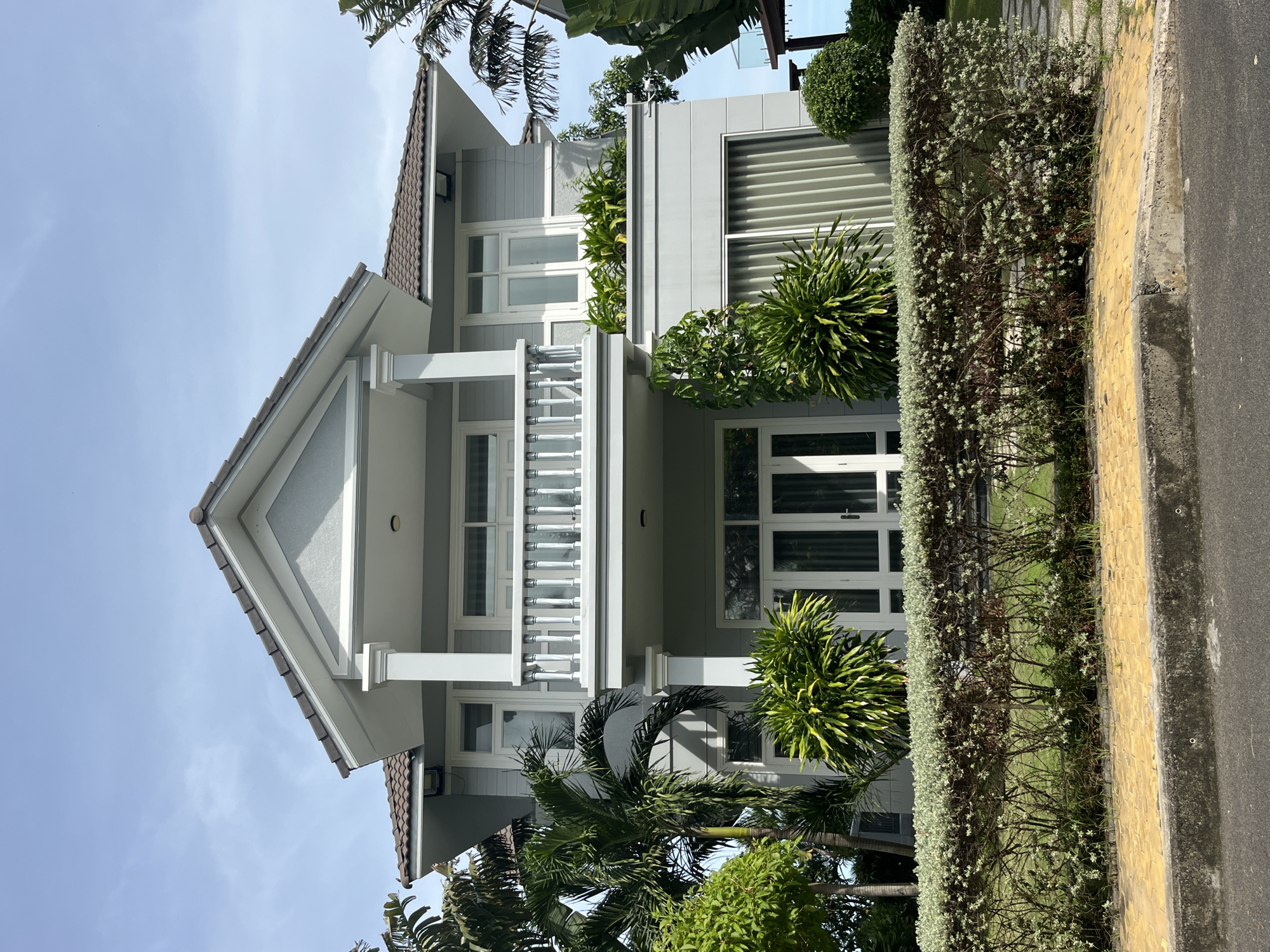 This is a frontal, eye-level, full shot of a modern two-story house, likely located in Phu Thuy Ward, Vietnam, given the tropical foliage. The house features a light grey facade with white trim around the windows and doors. The roof is tiled in a dark brown or grey color. A prominent feature is a balcony on the second floor, supported by white columns, with a white railing. On the ground floor, there are large double doors with glass panes. The front of the house is bordered by a neatly trimmed hedge with small white flowers, behind which lush green plants and palm trees are visible. A yellow paved walkway runs along the front of the hedge, separating it from the asphalt road. The sky is a clear blue with a few faint clouds, suggesting it is a sunny day. There are no people or animals visible in the image.