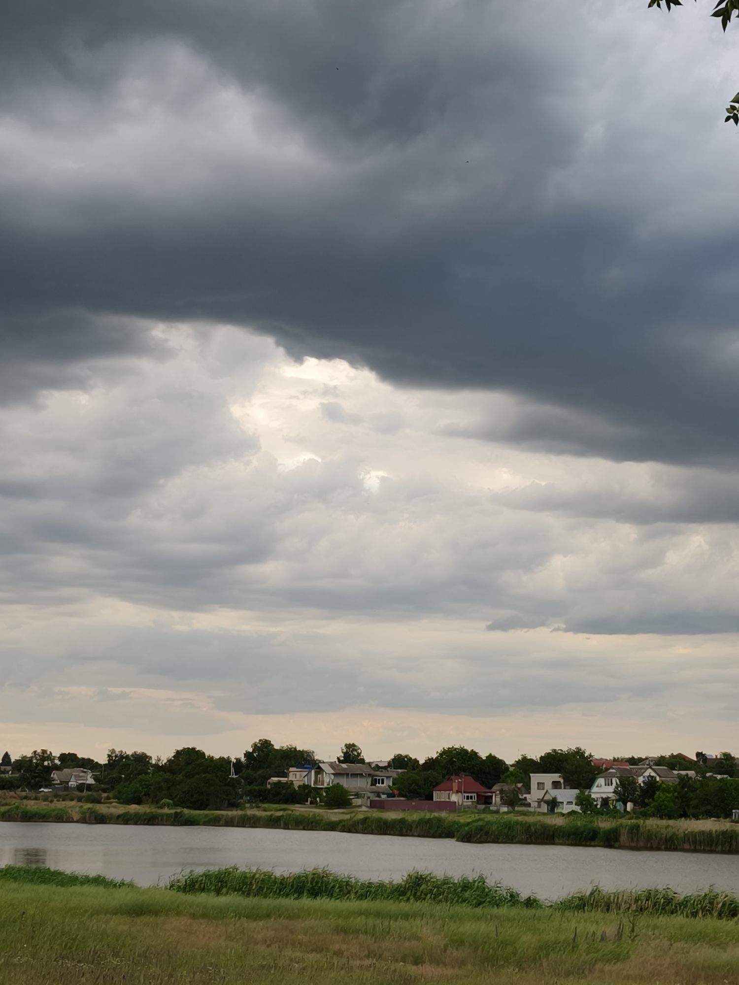 This is a vertical, medium-shot photograph of a rural landscape in Kryvyi Rih, Ukraine. The dominant feature is the dramatic sky, filled with thick, dark, grey storm clouds that stretch across the entire upper half of the image. A break in the clouds reveals a patch of brighter sky in the middle ground, suggesting diffused sunlight attempting to penetrate the gloom.
Below the sky, a body of water, likely a river or lake, forms the middle section of the frame. The water's surface is calm and reflects the muted tones of the sky. Along the far bank of the water, a small village or settlement is visible. Several houses with varied roof colors, including red and white, are nestled amongst dense green trees. The houses appear to be single-story residences, some with visible windows and chimneys.
In the foreground, a strip of tall, dry grass and reeds lines the edge of the water, giving way to a patch of green grass. The overall mood is somber and atmospheric, likely captured during a pre-storm period or shortly after a rain shower. The weather appears overcast and potentially windy, with the clouds suggesting an impending storm. There is no visible text in the image.