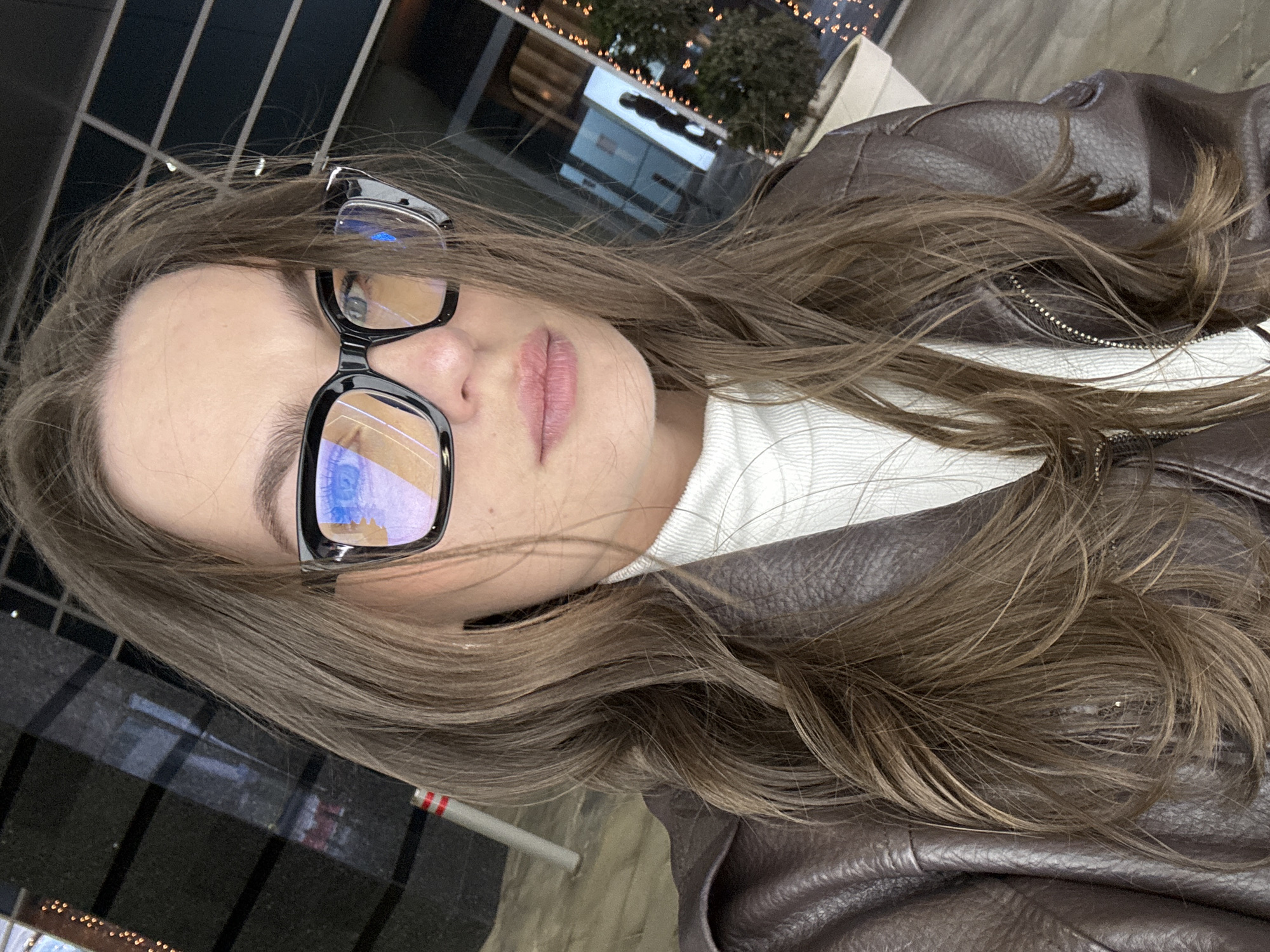The image features a woman wearing glasses and a brown leather jacket. She appears to be outdoors in an urban setting. Reflections in the glasses suggest a city environment with buildings. A possible cigarette is visible. It is not clear if the location is Moscow.