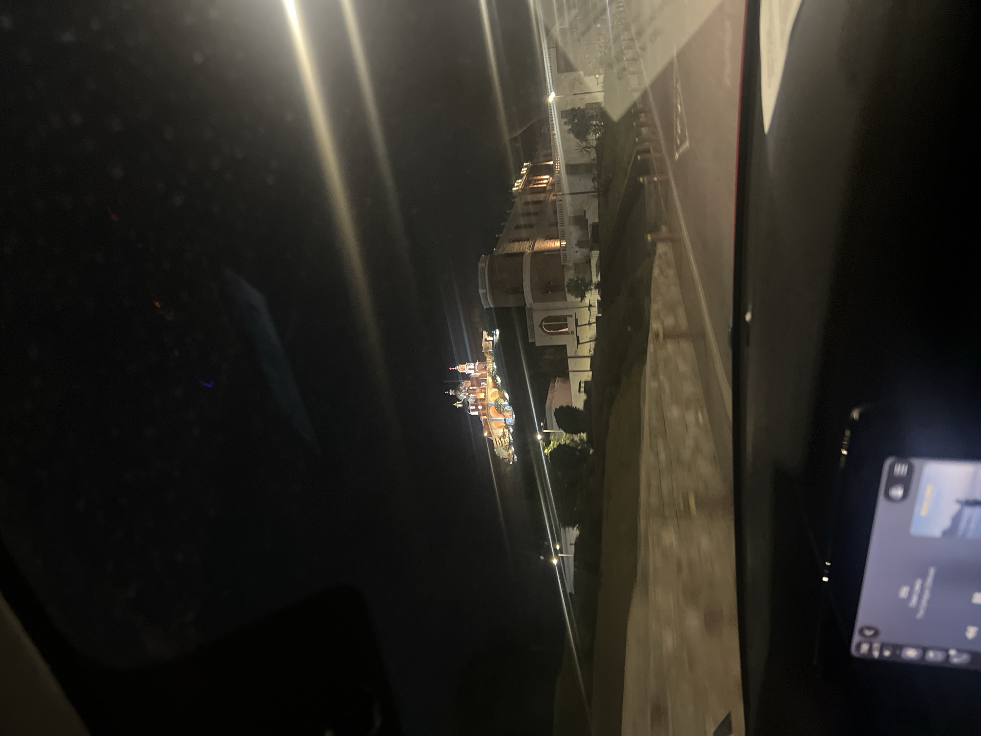 The photograph depicts a nighttime view of the city of San Andrés Cholula, Mexico, as seen from inside a vehicle. The illuminated Catedral de Nuestra Señora de los Remedios is visible in the distance. The image also captures other buildings and streetlights lining a road. A portion of the vehicle's interior, including a screen, is visible in the foreground.