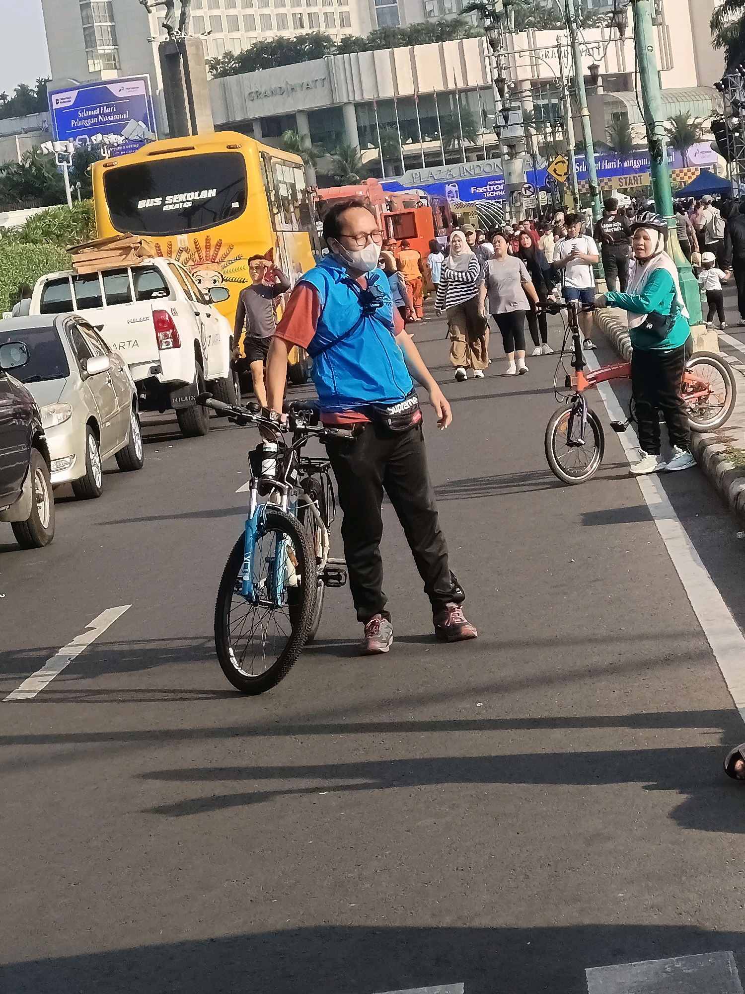 The image captures a street scene in Central Jakarta, Indonesia, likely during an outdoor event or organized activity. In the foreground, a man wearing glasses, a face mask, a red shirt, and a blue vest is standing next to his bicycle. The bicycle is black with blue accents. Behind him, a group of people are walking and cycling on the road, with several cars and a yellow school bus visible in the background. The buildings in the background suggest an urban setting, with one prominent building labeled "GRAND HYATT." Signs indicating "PLAZA INDONESIA" and "Selamat Hari Pelanggan Nasional" are also visible, hinting at a commercial or celebratory event. The road has white lane markings and shadows from overhead structures or trees are cast on the asphalt. The overall atmosphere appears to be one of public gathering and activity in a busy city environment.
