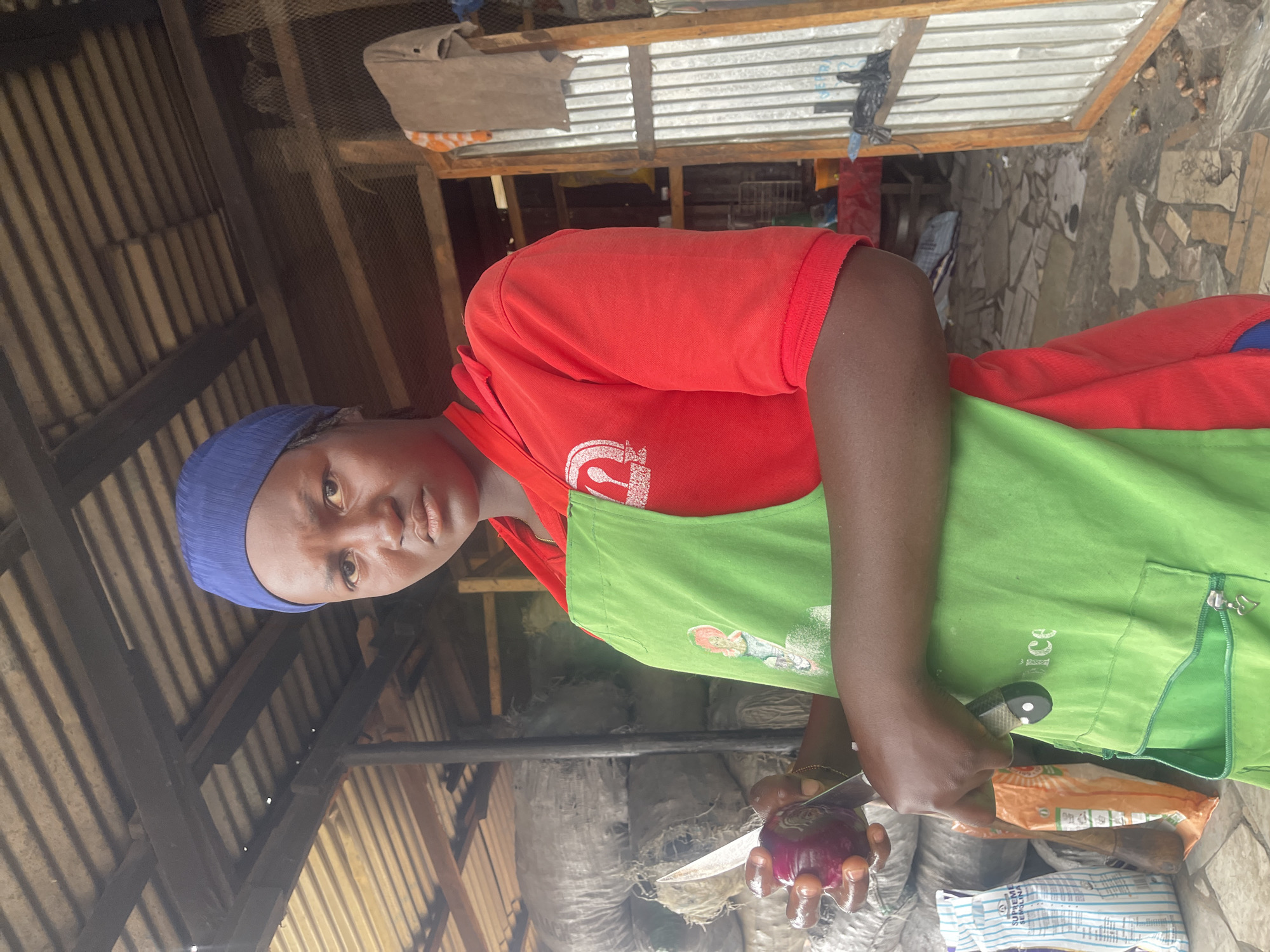 A person wearing a blue head covering and a red t-shirt with a green apron is holding a red onion and a knife. The person is located under a wooden structure with a corrugated metal roof in Jos, Nigeria. Bags of items are visible in the background.