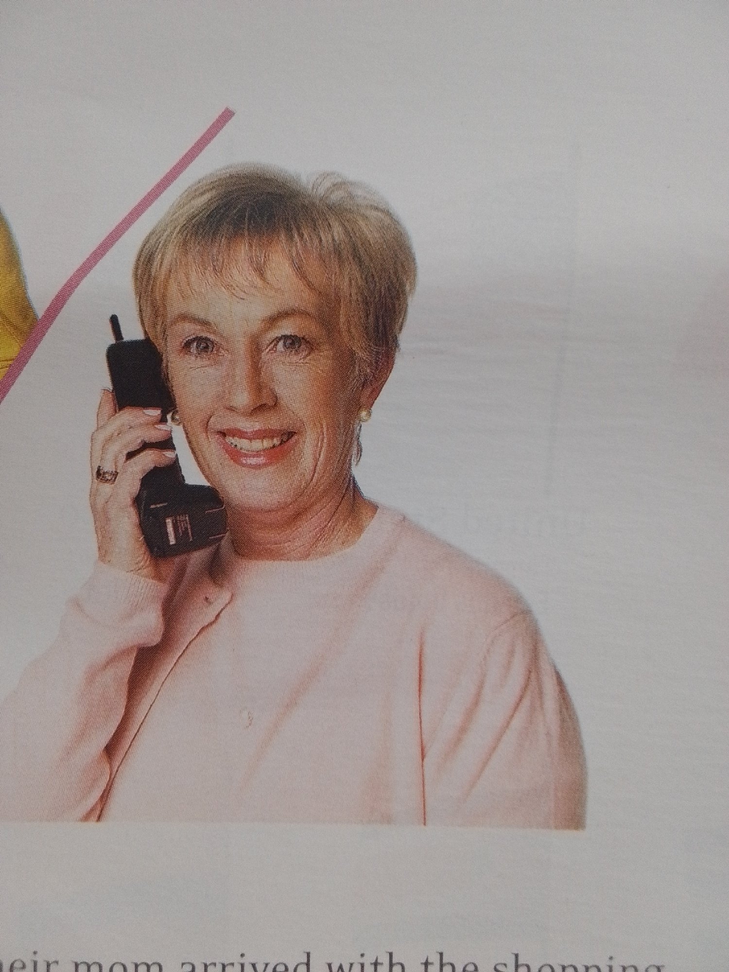 An elderly Caucasian woman with blonde hair is holding a black cordless phone to her right ear and speaking. She is smiling and wearing a light pink cardigan over a pink top. A ring is visible on her left ring finger. The background is plain white, with a diagonal pink stripe in the upper left corner. The woman is positioned in the center-right of the frame. The text "their mom arrived with the shopping" is visible at the bottom of the image, suggesting this is an illustration from a book or magazine. There is no indication of the location being La Tagua, Venezuela.
