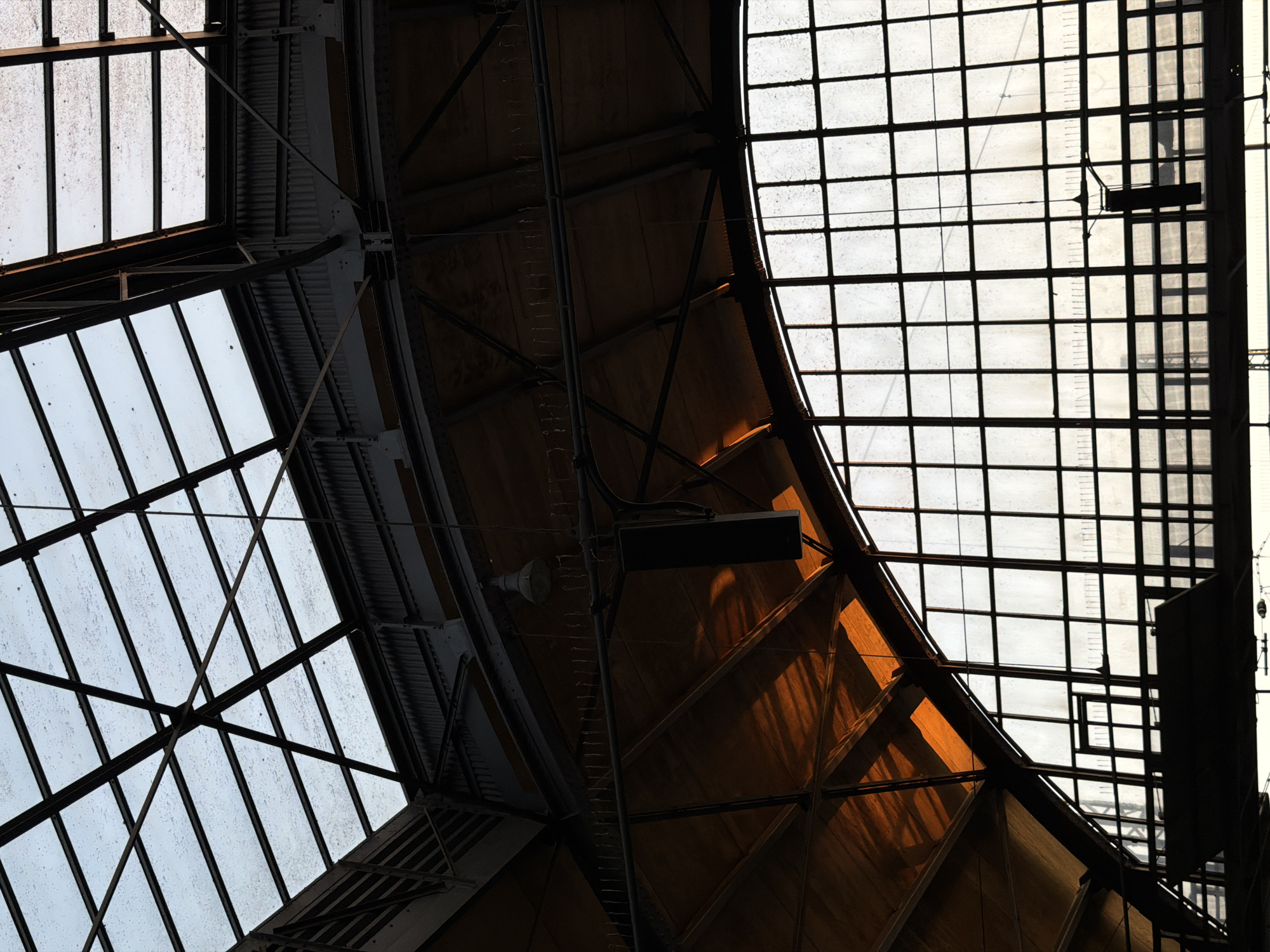 The image shows the interior of a large, industrial-style building with a high, arched ceiling made of glass and metal. The glass panels of the ceiling and the large, grid-like windows are partially obscured by dirt or condensation, creating a diffused light. Exposed metal beams and supports form a complex structure overhead. A large, orange-tinted panel, possibly part of the roof or an interior surface, is illuminated by light filtering through the glass. Hanging from the ceiling are what appear to be light fixtures or ventilation equipment. The overall impression is of a grand, somewhat weathered structure, possibly a train station or a market hall, bathed in the soft light of a cloudy or overcast day. This scene is located in the Krefeld Hauptbahnhof, Krefeld, Germany.