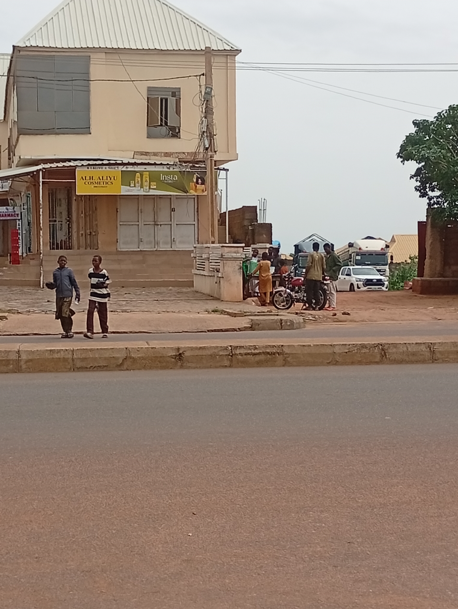 In Tumfure, Nigeria, a street scene unfolds with a building on the left featuring a light-colored facade and a corrugated metal roof. A sign above a storefront reads "ALH. ALIYU COSMETICS." Two young individuals are walking on a sidewalk in front of the building, one wearing a blue jacket and patterned pants, the other in a striped long-sleeved shirt and brown pants. Further to the right, a group of people are gathered near a red motorcycle and a white SUV. Several trucks are parked in the background. The environment suggests an urban or semi-urban setting with a road in the foreground and a sidewalk bordering the shops. The sky is overcast.