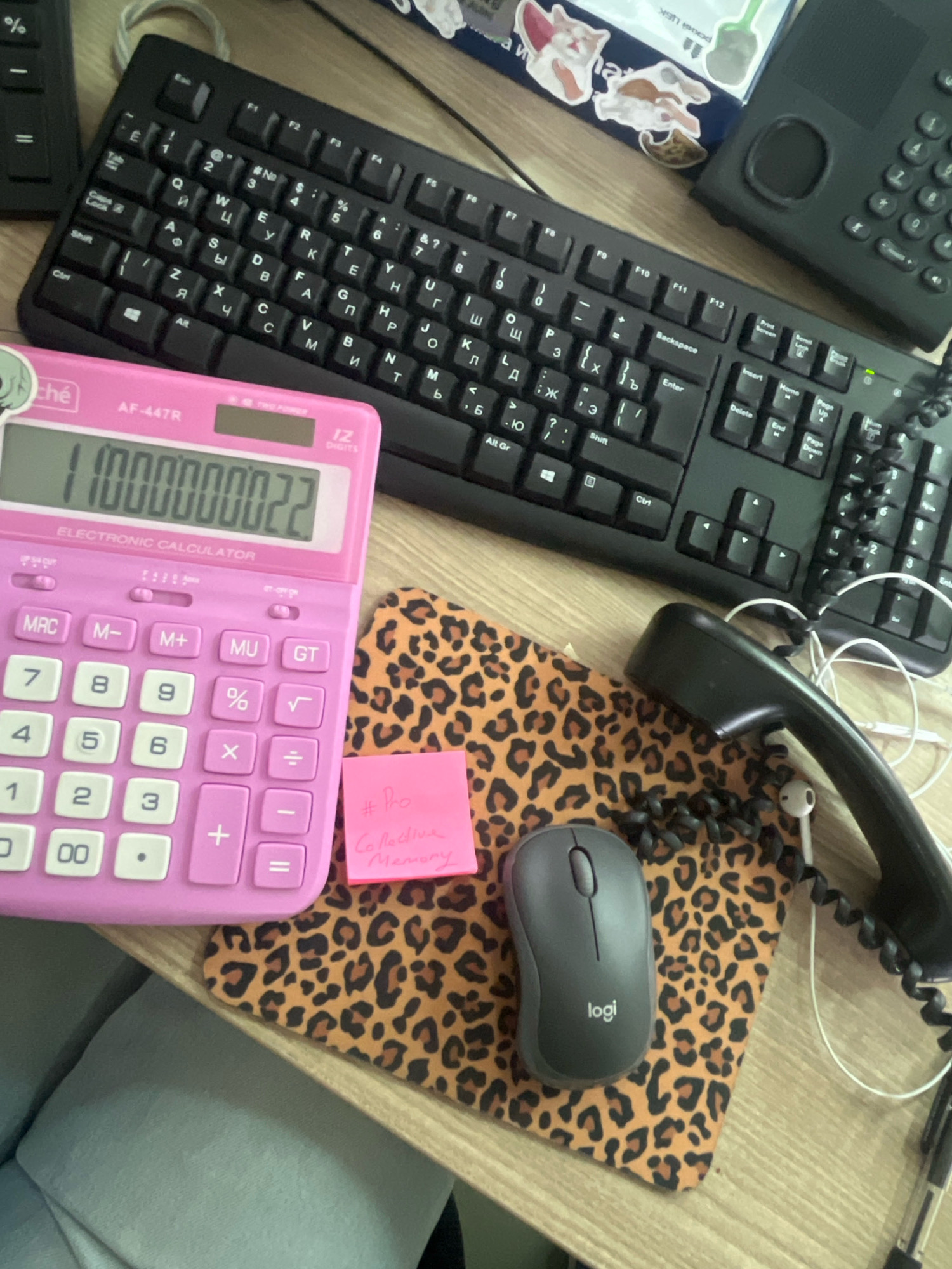 The image shows a desk with various office supplies. A black keyboard and a pink calculator are prominently displayed. Next to the calculator is a mousepad with a leopard print pattern. A wired telephone handset rests on the mousepad, along with a small pink sticky note. A dark gray computer mouse is also visible on the mousepad, with the "logi" logo clearly legible. In the background, part of a laptop and other electronic devices can be seen. The desk surface appears to be made of wood. The scene is indoors, likely an office or home workspace. There is no indication of the specific location in Moscow, Russia, or any actions or interactions occurring.