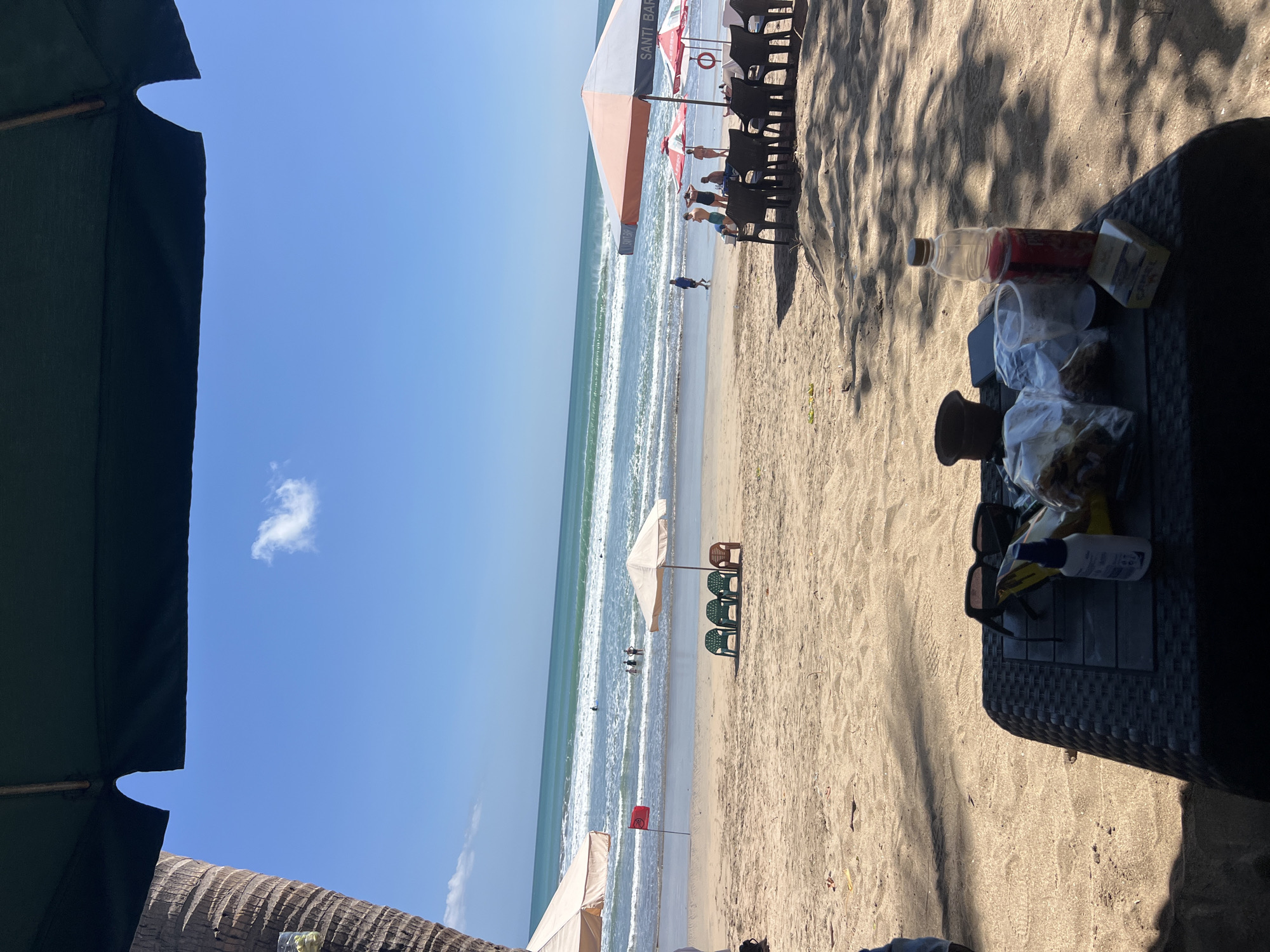 A wide-angle view captures a bustling beach scene in Kuta, Indonesia, under a clear blue sky. The foreground is dominated by a woven black table laden with various items: a bottle of red liquid, a pack of cigarettes, sunglasses, a small bottle of blue liquid, and plastic bags filled with food items. The table is positioned on the sandy beach, with dappled shadows from overhead umbrellas cast across it.
Beyond the table, rows of black plastic chairs and several beach umbrellas suggest a lively seating area. In the middle ground, a few empty green and brown plastic chairs are arranged near a white umbrella, with a red flag planted in the sand nearby. Further out, several people are visible wading in the ocean, enjoying the moderate waves. The turquoise water stretches to the horizon, meeting the clear blue sky. A tall palm tree trunk is partially visible on the left side of the frame, framing the upper portion of the scene along with the edge of a large, dark green umbrella. The sand itself is textured with footprints and small debris, indicating recent activity.