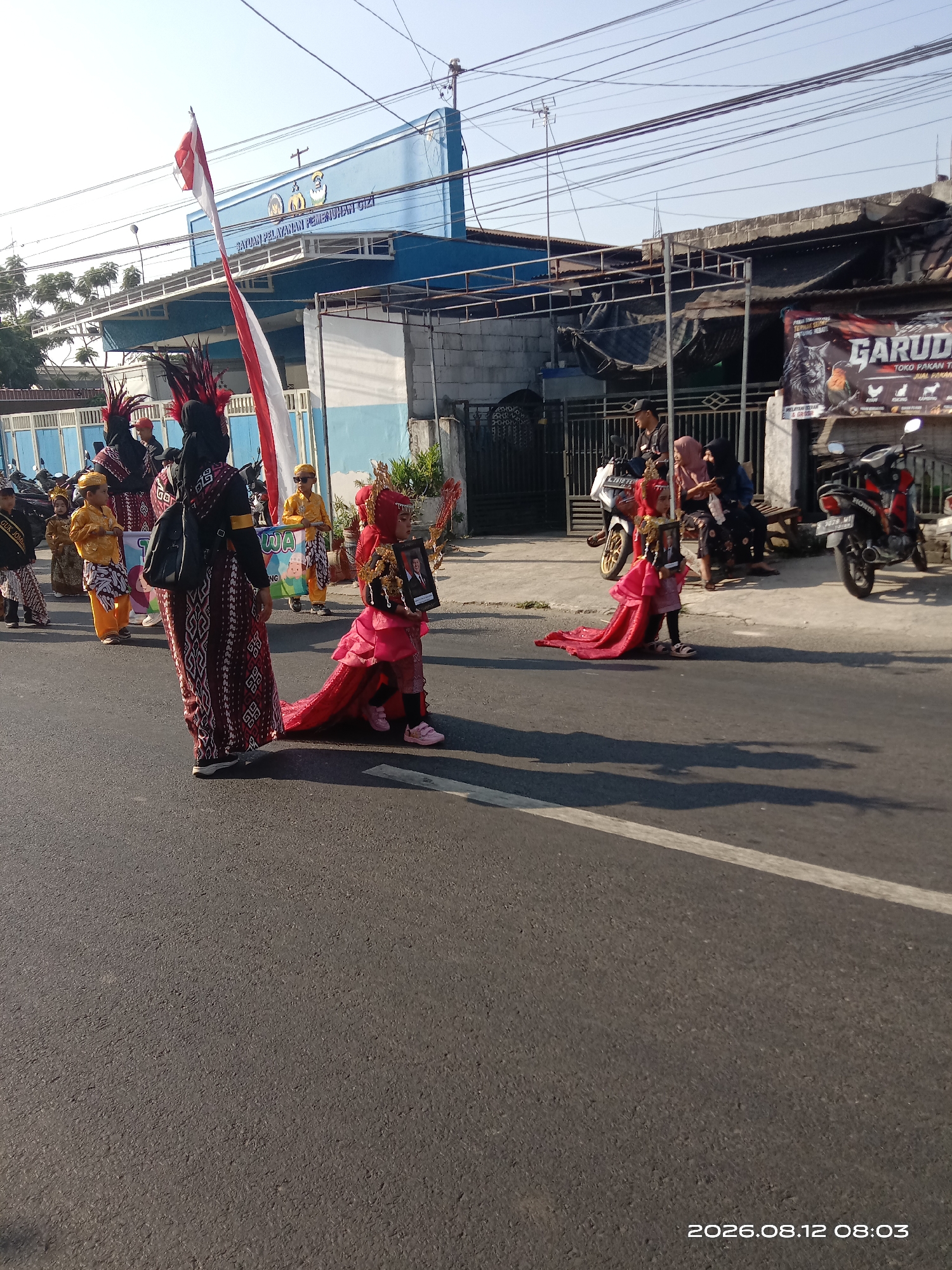 A parade is taking place on a street in Gudo, Indonesia. The image shows several people dressed in traditional attire, including children in colorful costumes with elaborate headdresses and long, flowing skirts. One child in the foreground is carrying a framed portrait of a man. Behind the paraders, a building with blue siding and white trim is visible, with a red and white Indonesian flag flying. A banner with the word "GARUDA" is displayed on a building to the right, along with images of animals. Several motorcycles are parked on the sidewalk. The sun is shining, casting shadows on the asphalt road. The timestamp "2026.08.12 08:03" is visible in the lower right corner of the image.