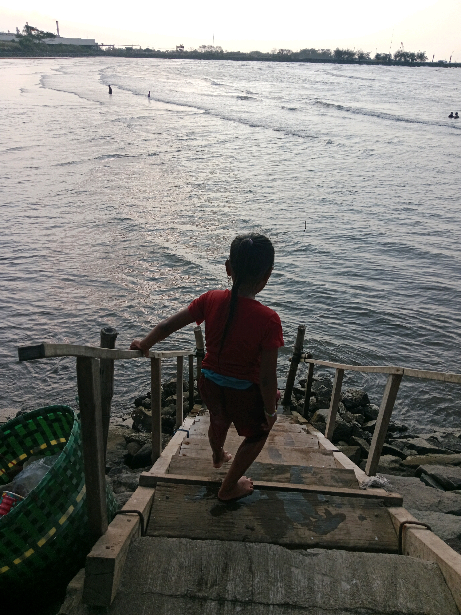 A young girl, seen from behind, stands on wooden steps leading down to the ocean. She has dark hair pulled into a braid and is wearing a red t-shirt and red shorts. The steps are weathered and appear to be part of a pier or boardwalk structure. To the left, a large green woven basket with some items inside is visible. The ocean is calm with small waves, and in the distance, several people can be seen in the water and on the shore. The background shows a coastline with vegetation and structures, possibly industrial buildings. The lighting suggests it is late afternoon or early evening. The scene is located at the coast of Probolinggo, Indonesia.