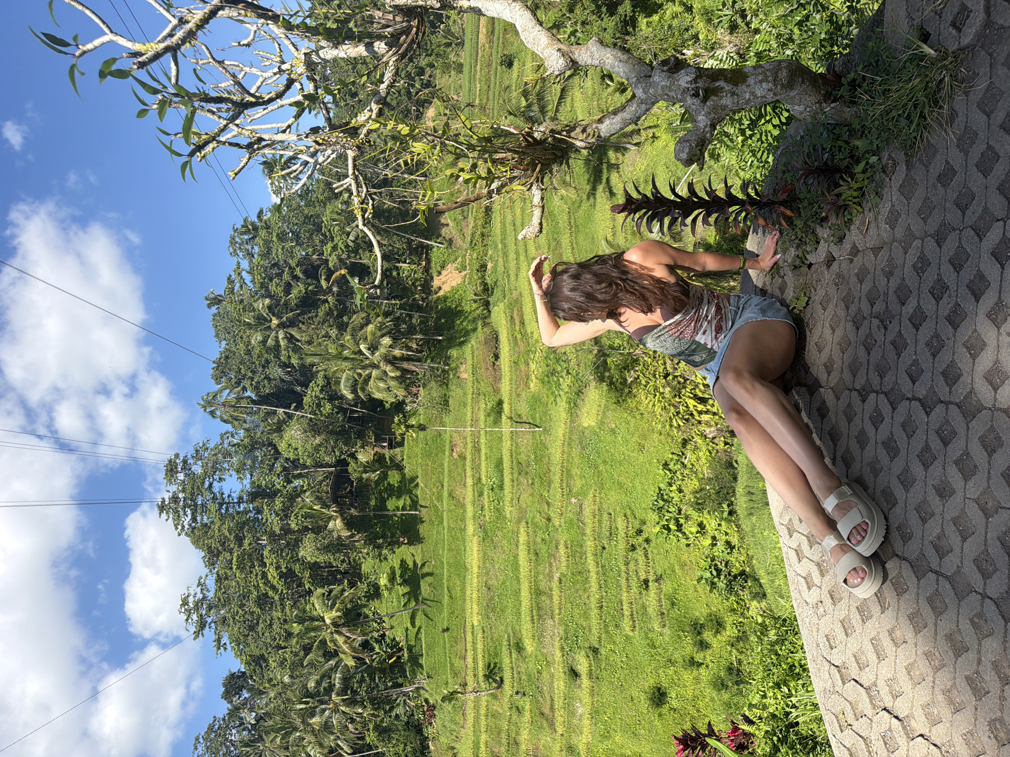 In Tegallalang, Indonesia, a woman sits on a patterned stone surface, facing away from the camera. She has long, brown hair and is wearing a patterned scarf as a top and denim shorts. Her left arm is raised, with her hand resting on her head. Her legs are extended forward, and she is wearing light-colored platform sandals. In the background, lush green rice terraces descend down a hillside, dotted with palm trees and other tropical vegetation. The sky is a vibrant blue with scattered white clouds. Several power lines are visible stretching across the frame above the trees. To the right, a gnarled tree with sparse leaves stands prominently in the foreground.