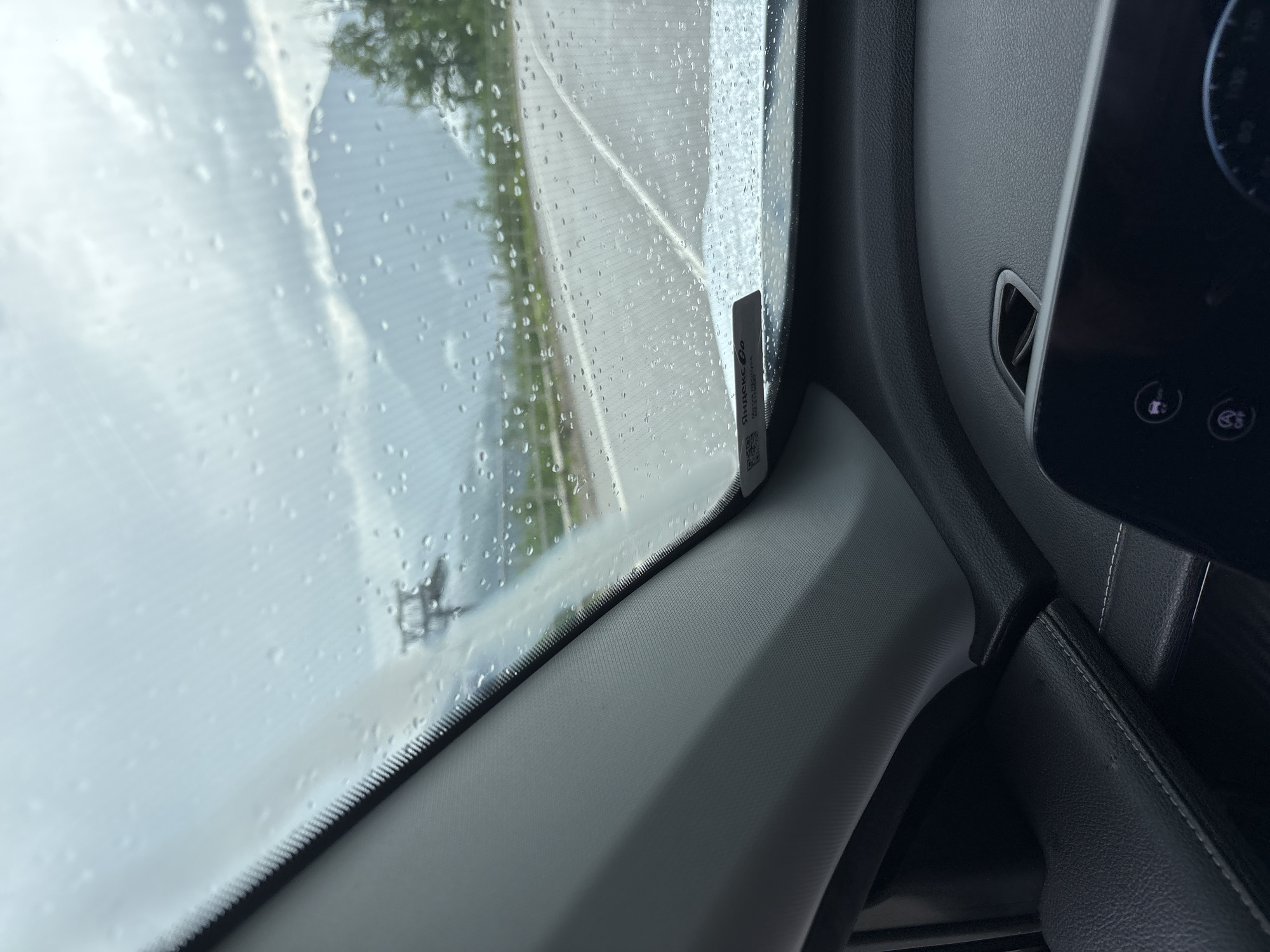 The image shows a view from inside a car in Estosadok, Russia, on a rainy day. Water droplets are visible on the windshield, obscuring the view of the road, mountains, and trees outside. The interior of the car, including the dashboard and a portion of the infotainment screen, is in the foreground. A sticker with the "Yandex Go" logo is affixed to the windshield. The weather appears overcast and the overall atmosphere is damp and subdued.