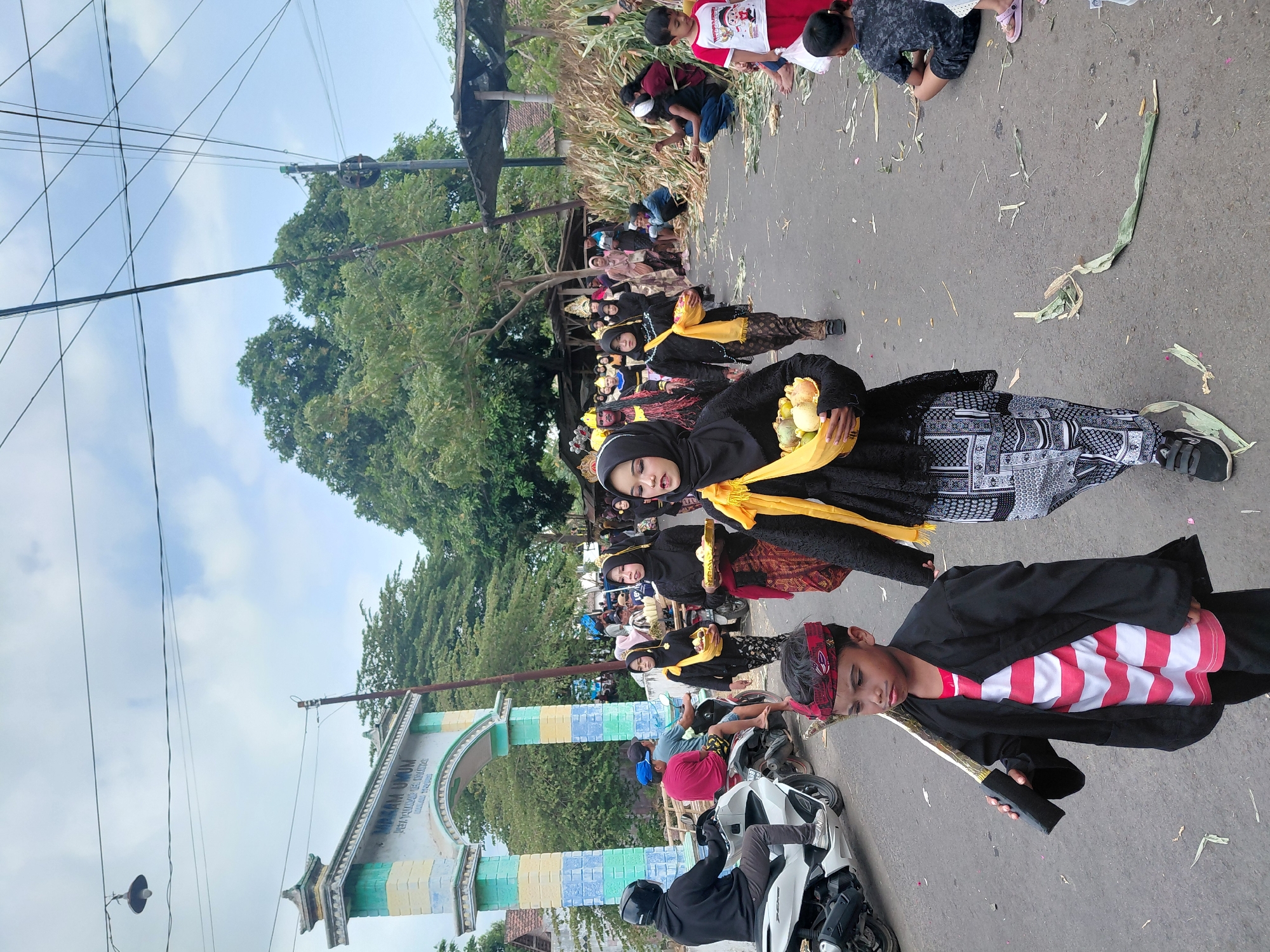 A group of people, some in traditional attire, are participating in a procession or parade on a paved road. The group includes children and adults, with some women wearing black outfits and headscarves, adorned with yellow sashes and carrying bowls of fruit. A young boy in the foreground wears a black jacket over a striped shirt and carries a prop sword. In the background, there is an archway with text that appears to read "MAKAM UMUM" and "DESA TAMBAKSARI". Other people are visible watching the procession from the side of the road, where piles of dried corn stalks are stacked. A white motorcycle is parked on the left side of the road. The scene takes place outdoors under a cloudy sky with power lines visible overhead. The location is identified as Bimo, Indonesia.