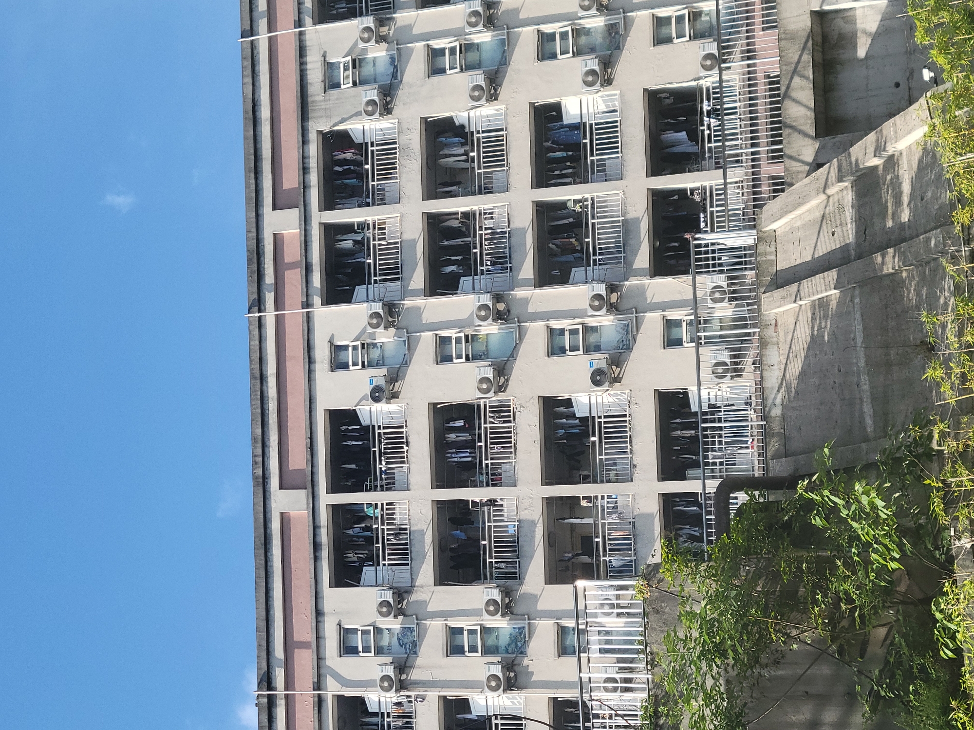 An upward shot of a building with multiple windows and air conditioning units in Beibei, China, presumably an apartment complex. Clothes are hanging inside many of the units. The building is next to a tall concrete wall covered with green plants. The sky is blue and clear.