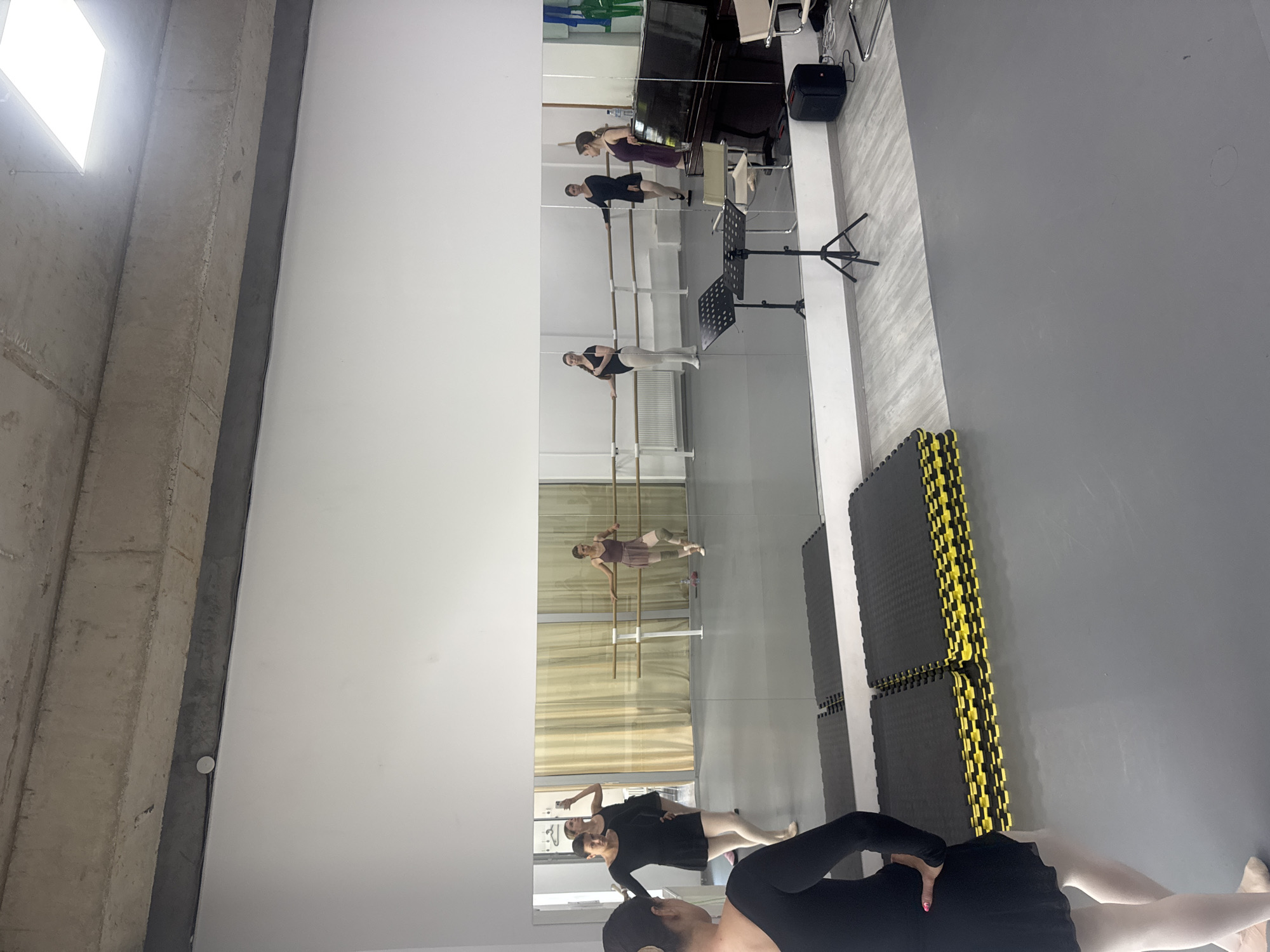 The image depicts a ballet studio in Tbilisi, Georgia. A group of ballerinas are practicing at the barre, visible in the mirrored wall. The studio features a gray floor partially covered with black and yellow mats. A piano, music stand, and other equipment are reflected in the mirror. The ceiling is white with a rectangular light fixture. The walls are white and gray concrete.