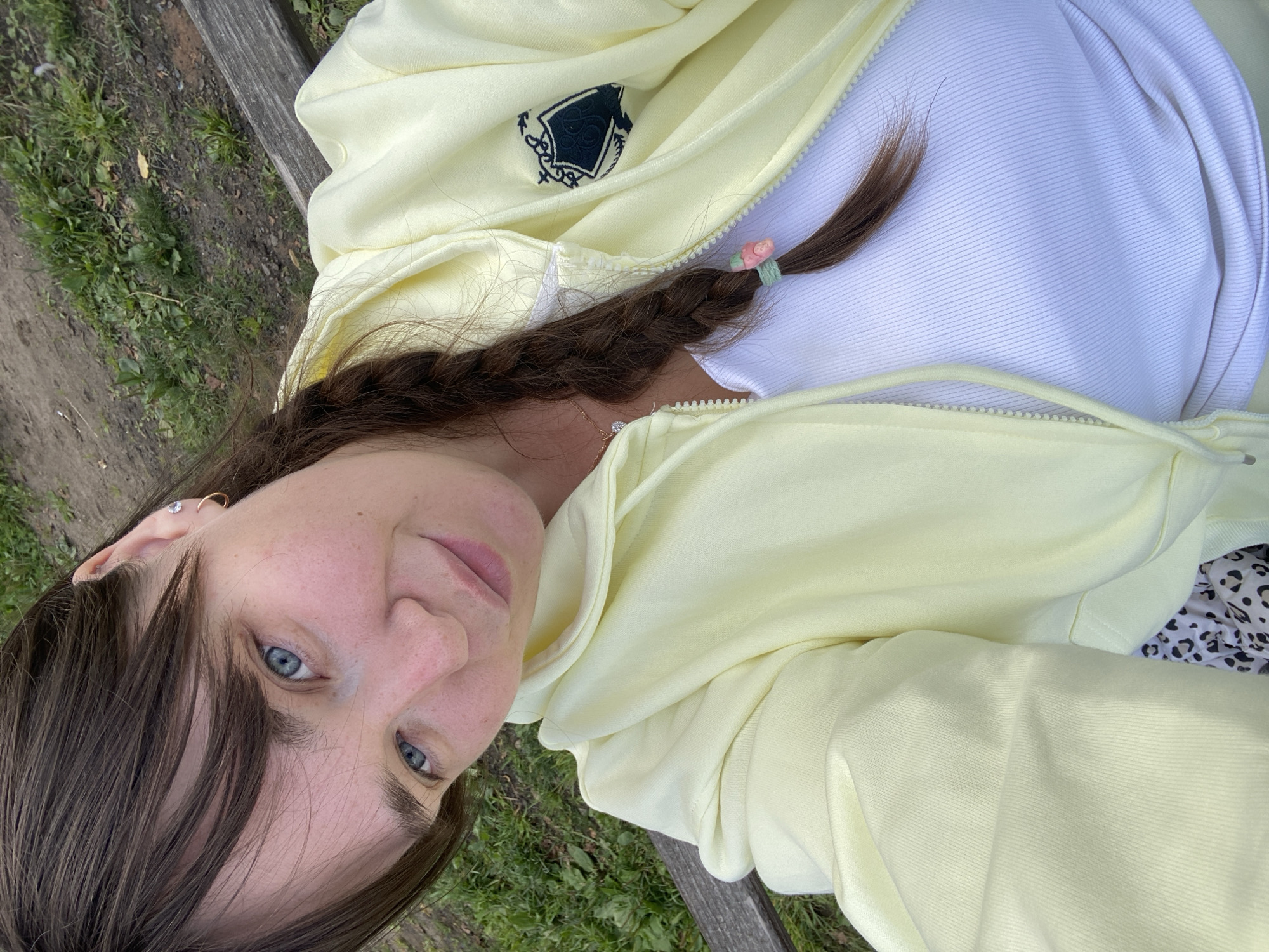 The image shows a close-up, high-angle selfie of a woman sitting on a park bench. The woman has brown hair in a braid, blue eyes, and fair skin. She is wearing a light yellow zip-up hoodie over a white ribbed tank top and has a small necklace on. A small patch with a crest is visible on the left side of the hoodie. The background consists of a dirt path and green grass. The scene appears to be an outdoor public space, likely a park, in Votkinsk, Russia, given the requested location.