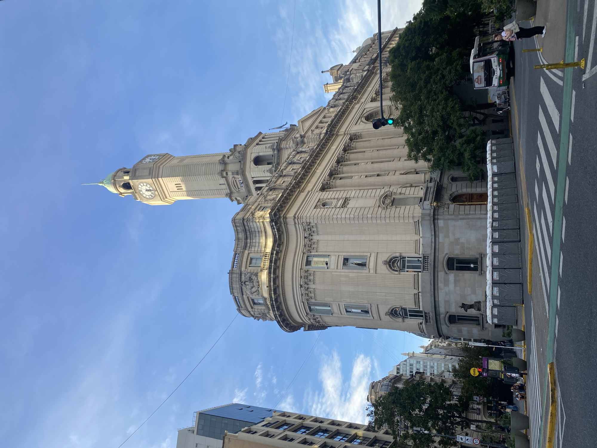 The image shows Palacio de Correos in Buenos Aires, Argentina. The building is tall, made of light-colored stone, and has a clock tower. The sky is blue with some clouds. There are some modern buildings visible. A bus and people are present on the street with some mobile toilets placed in a row along the building. A traffic light is visible with a green light.