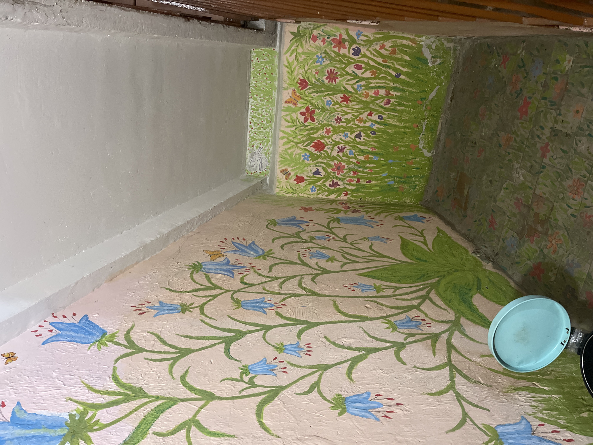 The image shows a corner of what appears to be an indoor stairway with painted murals on the walls and floor. The left wall is painted with large blue bell-shaped flowers and green stems against a light pink textured background. Several small orange butterflies are also depicted on this wall. The floor is painted with a floral pattern in shades of green, pink, and orange. The right wall features a mural of a green grassy field with various small colorful flowers, including red, pink, blue, and yellow, and two orange butterflies. Above this mural, on a narrow ledge, a smaller mural depicts a white rabbit-like creature in green grass. The ceiling is white and slopes downwards towards the right. A light blue trash can lid is visible on the floor near the left wall. A wooden railing is partially visible on the far right side of the image. The scene is dimly lit, suggesting an indoor environment.