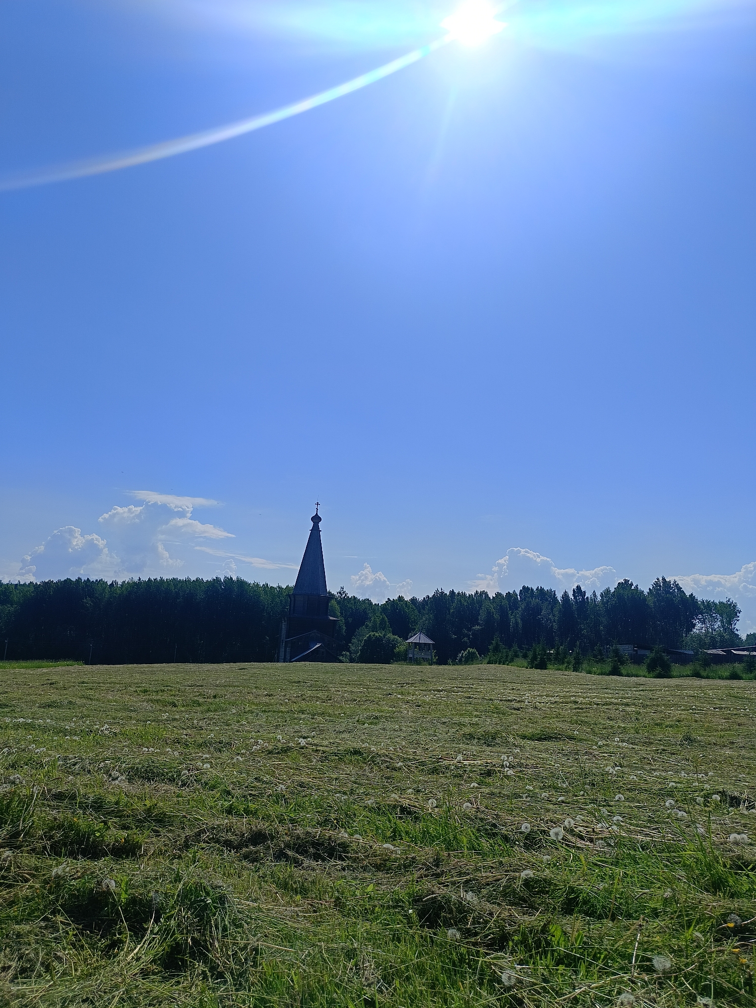 A sun flare from the upper right corner of the frame illuminates a bright blue sky. In the middle ground, a dark, wooden, steeple-topped structure, possibly a church, stands to the left of a small gazebo and is surrounded by a dense line of dark green trees. In the foreground, a field of freshly cut grass, with some dandelions scattered throughout, stretches across the bottom of the image. The scene is a rural landscape with a building and a field under a clear sky. The location is the village of Popovkа, Russia.