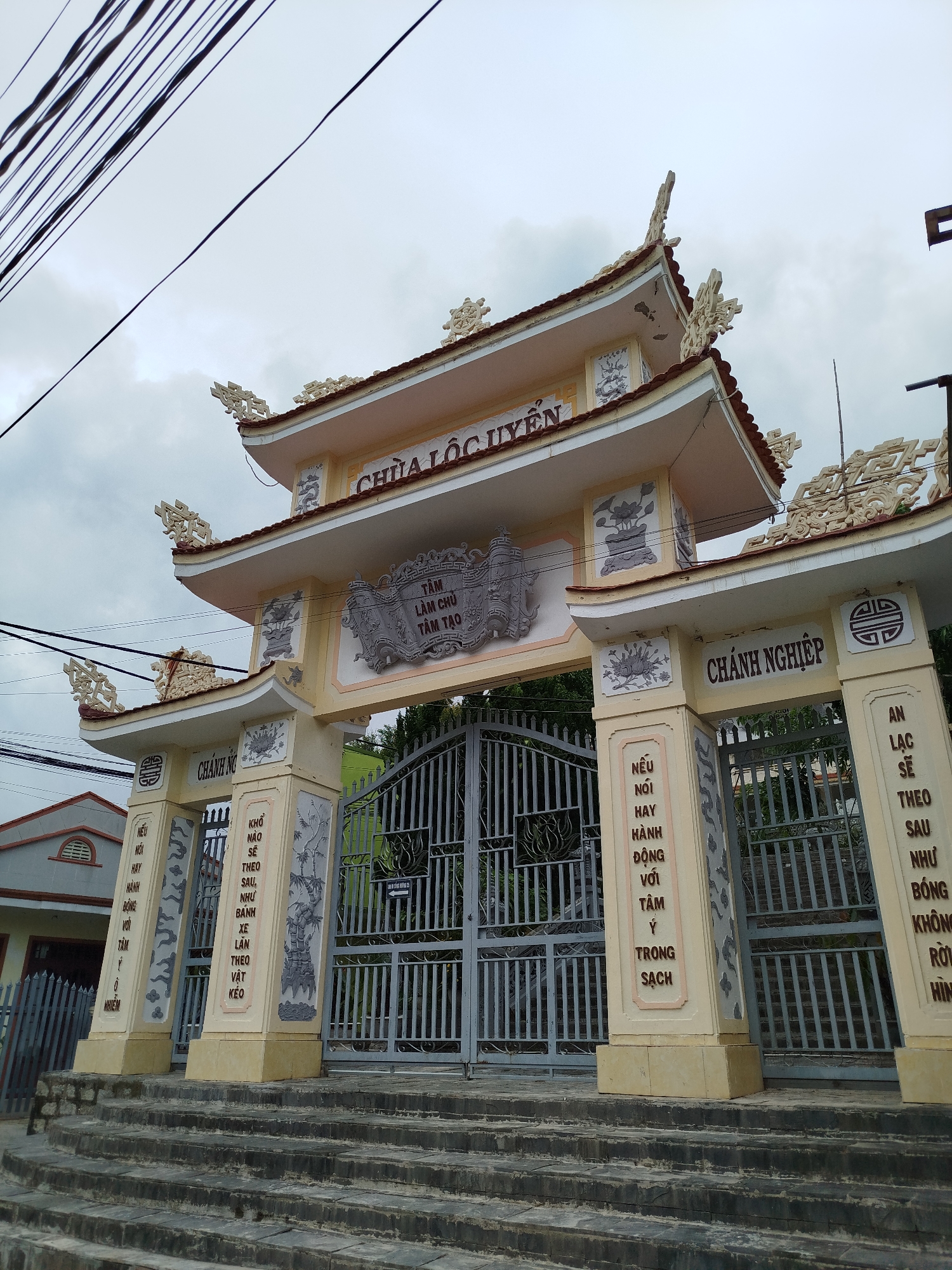 The image displays the ornate entrance gate of Chùa Lộc Uyển (Lộc Uyển Pagoda) in Da Lat, Vietnam, under an overcast sky. The gate is an elaborate structure built in a traditional Vietnamese architectural style, featuring multiple tiered roofs with decorative, upward-curving eaves adorned with intricate white sculptural elements. The main body of the gate is a warm cream color, complemented by grey accents and decorative panels, some depicting lotus flowers.
A series of dark, weathered stone steps leads up to the gate. The actual entrance is secured by large, grey metal gates, which are currently closed and feature a stylized lotus flower design. Above these gates, a prominent central plaque displays the Vietnamese text "TÂM LÀM CHỦ TÂM TẠO," translating to "Mind is master, mind creates."
On the left main pillar, beneath the heading "CHÁNH NG" (likely an abbreviation related to Buddhist principles), the visible text reads: "NẾU NÓI HAY HÀNH ĐỘNG VỚI TÂM Ý TRONG SẠCH" (If one speaks or acts with a pure mind/intention). On the inner face of this pillar, another Buddhist quote states: "KHỔ NÀO SẼ THEO SAU, NHƯ BÁNH XE LĂN THEO VẬT KÉO" (Suffering will follow, like the wheel of a cart following the ox that pulls it).
The right main pillar, headed "CHÁNH NGHIỆP" (Right Livelihood/Action), carries the inscription: "AN LẠC SẼ THEO SAU NHƯ BÓNG KHÔNG RỜI HÌNH" (Peace and happiness will follow like a shadow that does not leave the body).
No people are visible, lending a quiet and serene atmosphere to the scene. In the upper left corner, several black electrical wires crisscross the cloudy sky. To the left of the gate, a portion of a residential building with a grey roof is visible.