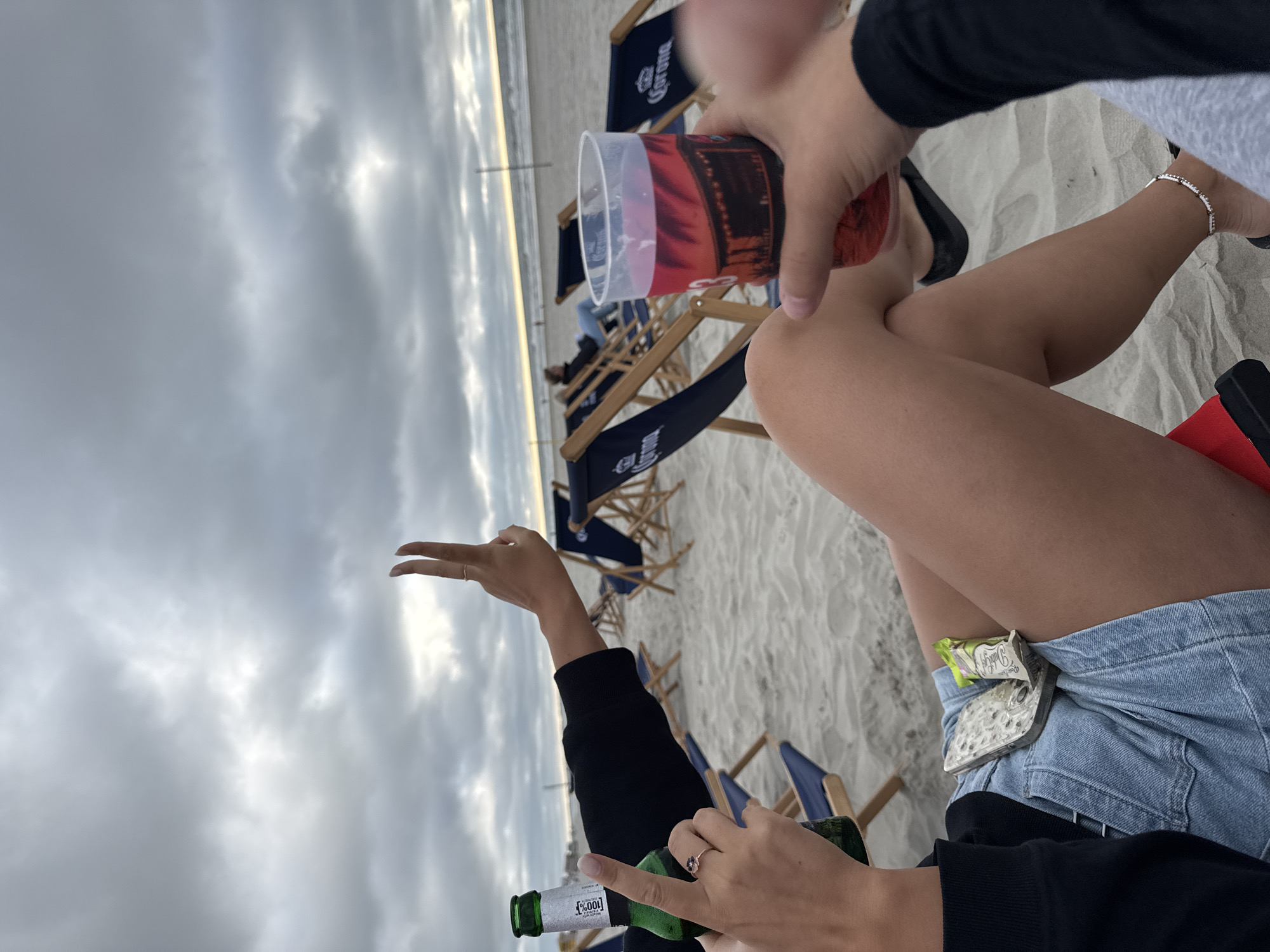 Two individuals are on a sandy beach, facing the ocean. The sky is overcast. In the foreground, one person holds a green beer bottle and shows two fingers extended. To their right, another person holds a red drink in a clear plastic cup. Both individuals are seated in beach chairs, with several other empty beach chairs visible in the background. The scene suggests a relaxed atmosphere on a beach, possibly during a cloudy day in Langeoog, Germany.