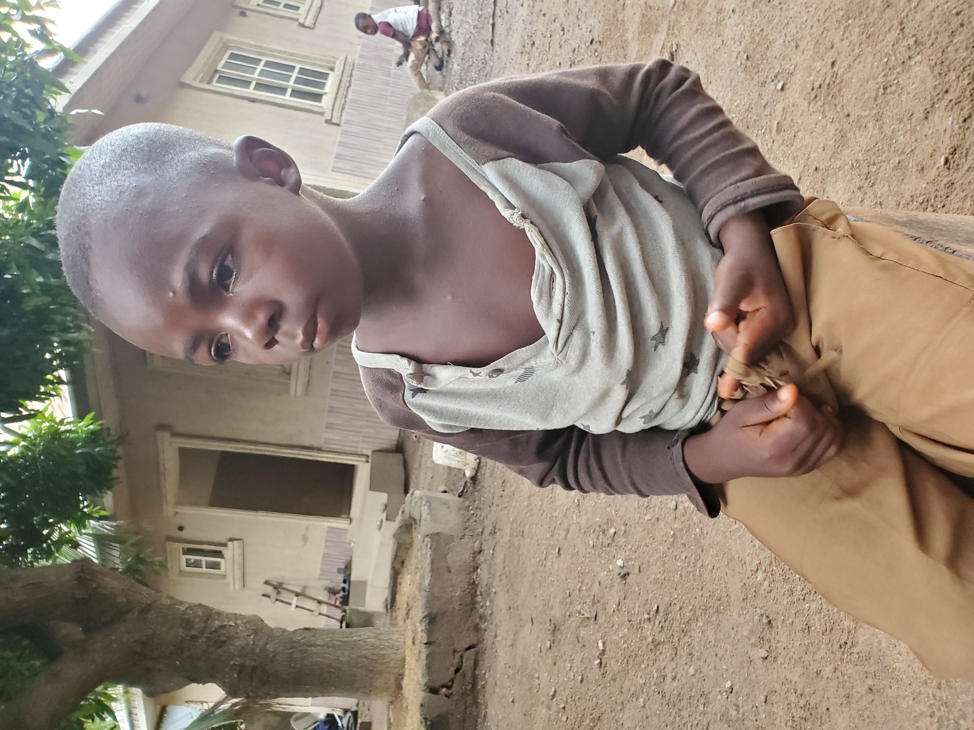 A young, dark-skinned child with a shaved head is standing outside, holding tan-colored cloth or pants in their hands. The child is wearing a brown long-sleeved shirt and a gray tank top with star patterns. The background includes a light-colored building with windows, a tree, and an unpaved ground area. Another person is faintly visible further back near the building. The city is Unknown, Unknown.