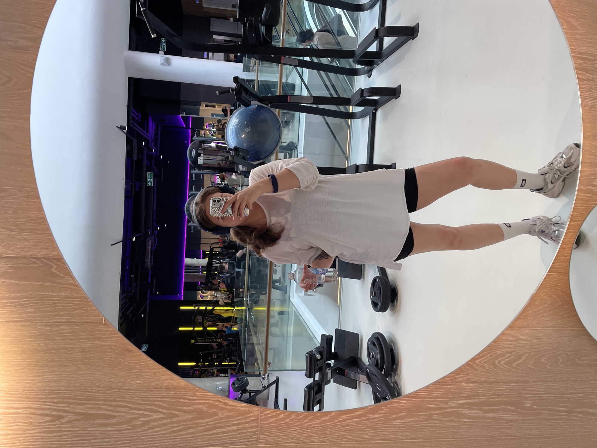 The image depicts a mirror selfie taken by a female individual within a modern, multi-level fitness club, specifically identified as a World Class facility located in Kuntsevo Plaza, Moscow, Russia.
The primary subject stands facing the camera's reflection in an oval, light-wood-framed mirror. Her face is partially obscured by a smartphone in a striped protective case. She wears wireless over-ear headphones, a loose white long-sleeve top, black athletic shorts, white socks, and light-colored athletic shoes. A blue wristband is visible on her left arm, and she holds a clear plastic water bottle with a blue label in her right hand.
The reflected environment reveals a spacious, brightly lit gym floor contrasting with a dark ceiling adorned with prominent purple and vertical yellow LED strip lighting. Various pieces of black fitness equipment are visible, including weight plates on the floor, a hyperextension bench, and a blue balance trainer. The gym's multi-level structure is evident through glass railings separating floors and visible escalators in the background. Several blurred individuals are present in the background, indicating an active gym setting. Green exit signs with Cyrillic text are mounted on the walls.