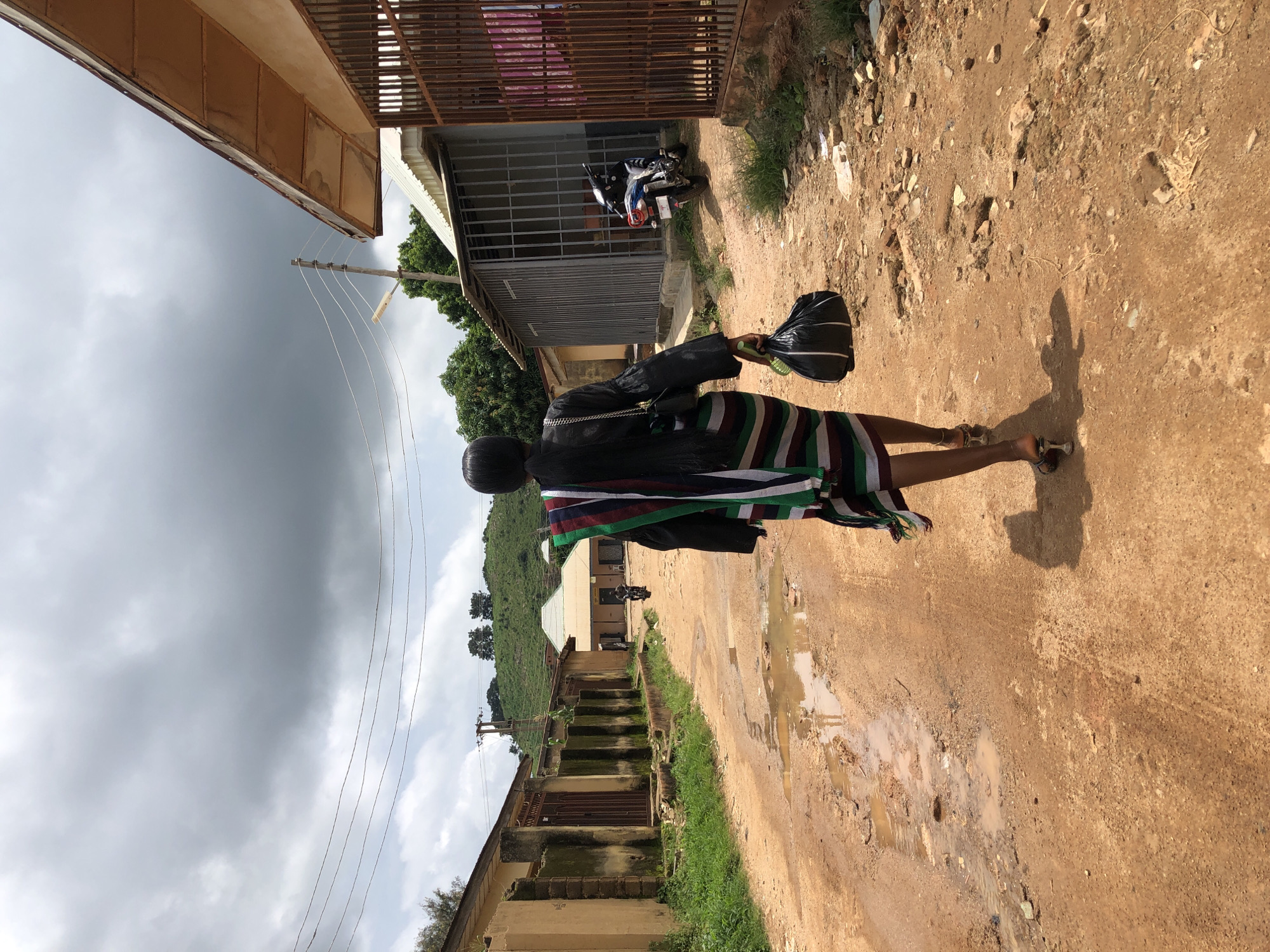A young woman walks away from the camera down a muddy, unpaved road in Jos, Nigeria. She is wearing a black ruffled jacket over a striped skirt and high heels, carrying a black plastic bag in her right hand. To her left, a row of tan buildings with metal gates line the road, some with overgrown grass and moss. To her right, a building with a corrugated metal roof and barred windows is visible, with a blue motorcycle parked in front of it. In the distance, more buildings and a green, hilly landscape are visible under a cloudy sky.