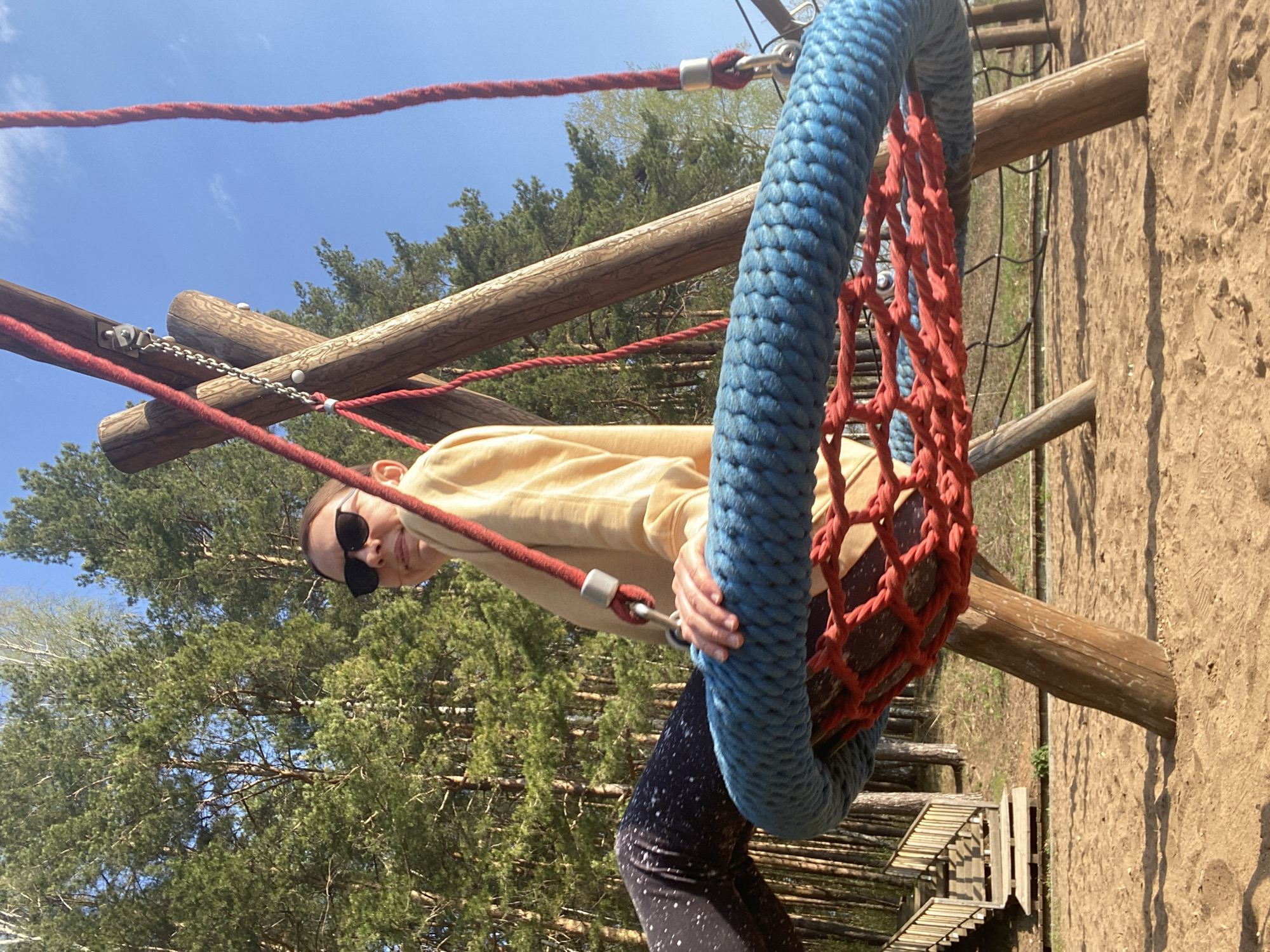 The photograph depicts a woman on a playground structure in Chaikovsky, Russia. The woman is wearing sunglasses, a light-colored long-sleeved shirt, and dark leggings with light-colored speckles. She is holding onto a swing made of a blue rope outer ring and a red rope net inside. The swing is suspended from a wooden structure with red ropes. The background consists of green trees and a blue sky. To the right, there's a sandy area and a wooden structure that resembles a bridge or walkway. The overall scene appears to be a sunny day in a park or recreational area.
