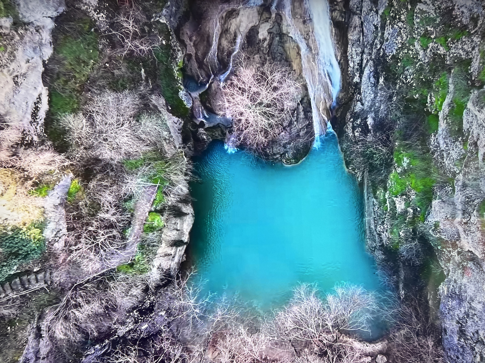 The image is an aerial view of a body of water, likely a pool or small lake, with a vibrant turquoise color. Waterfalls cascade into the pool from above, and the surrounding landscape is characterized by rocky cliffs with patches of green vegetation, possibly moss or grass. Leafless trees or shrubs are clustered around the edges of the pool and along the cliffs, suggesting it may be wintertime. In the bottom left, a set of stone steps is visible, adding an element of human influence to the scene.
While the image is reminiscent of several locations with waterfalls and rock formations, there are no recognizable features to confirm the location is in Basingstoke and Deane, United Kingdom.