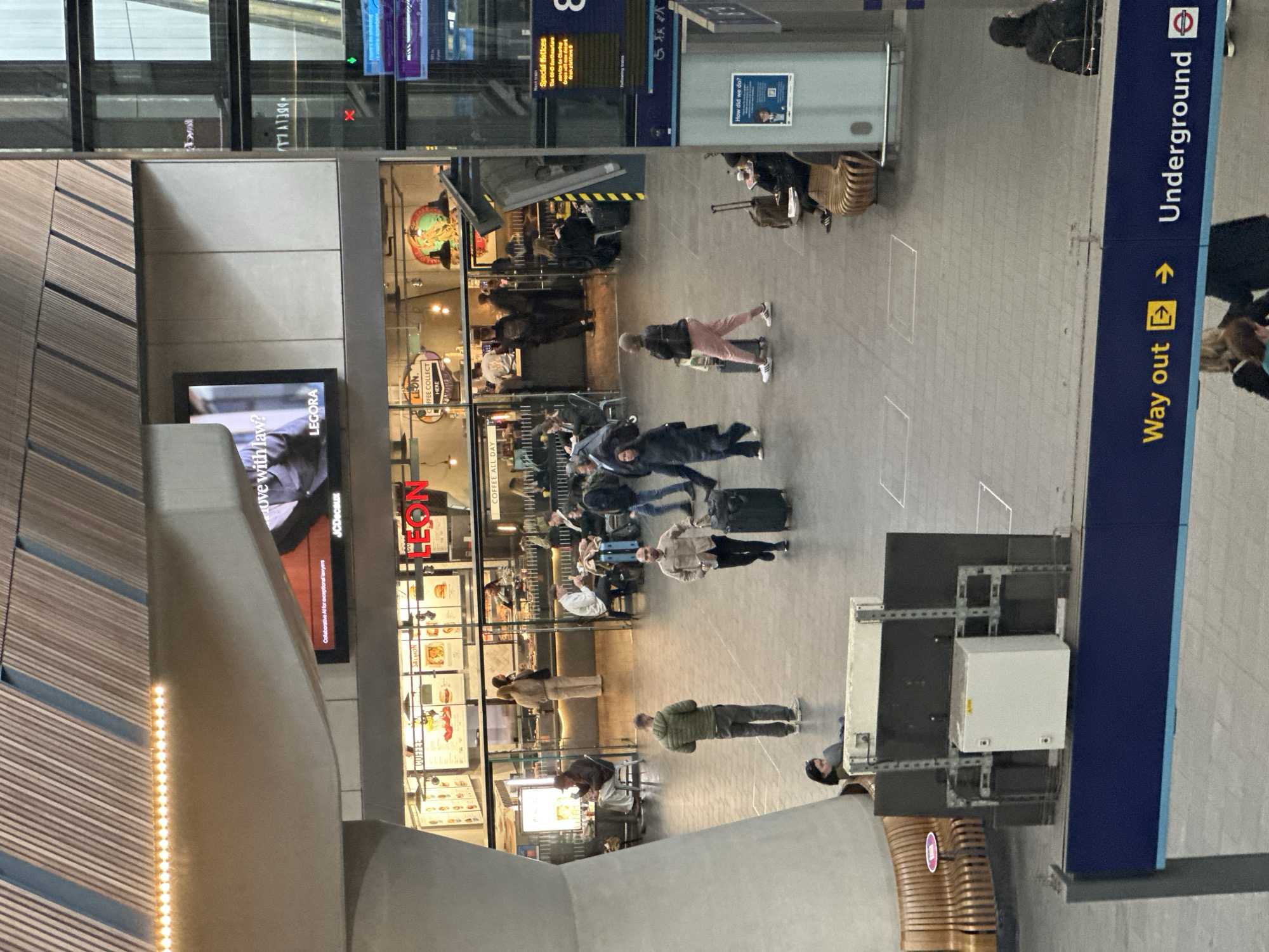 Here is a description of the image:
The image depicts a bustling scene inside a train station in London, United Kingdom. The focus is on a wide, open corridor where people are walking with their luggage.
To the right, a sign points towards the "Underground," indicating the location is a London Underground station. There are directional signs.
The area includes a branch of LEON, a fast-food restaurant chain, with its menu displays visible behind a glass facade. Passengers are seen browsing the menu.
A large advertising screen is also visible to the left.