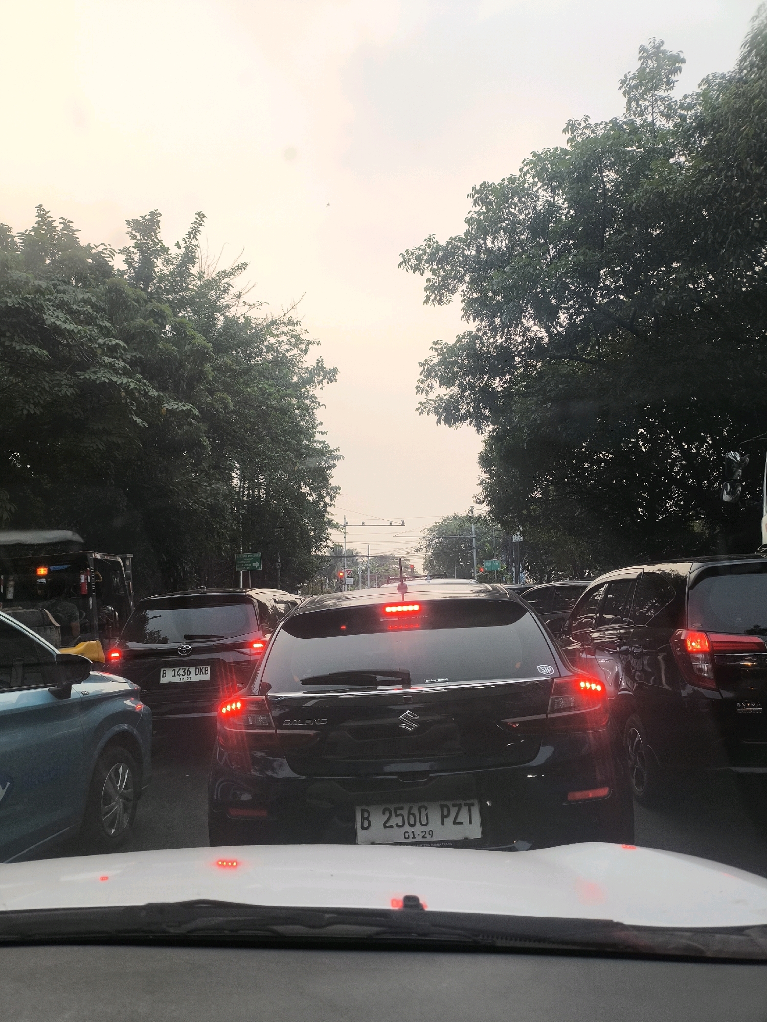 The image depicts a traffic jam on a street in South Jakarta, Indonesia, as indicated by the vehicle license plates, which start with "B" and appear to follow Indonesian formatting. The perspective is from inside a car, looking forward. Several vehicles are visible, including a blue car in the foreground to the left, a black Suzuki Baleno in the center with its brake lights illuminated, and other cars partially obscured. Trees line both sides of the street, creating a canopy overhead, and the sky is overcast with a hazy, light-colored atmosphere, suggesting either early morning or late afternoon. Traffic signals and street signs are also visible in the distance. The overall scene is typical of urban traffic congestion.