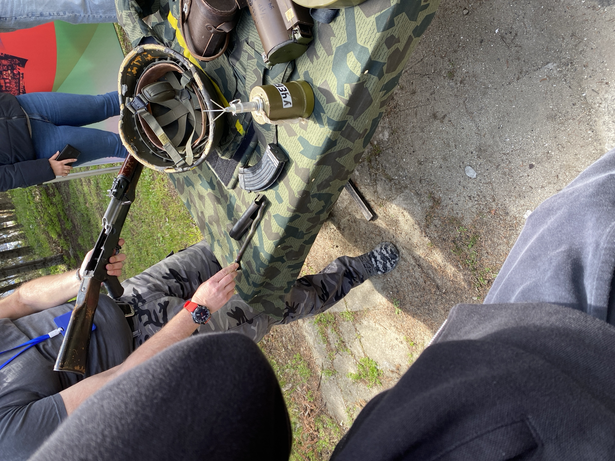 The image, captured from an elevated perspective looking down, depicts an outdoor military-themed display or event, likely in Varna, Bulgaria.
In the center, a table draped with a green and brown camouflage cloth showcases various military-style items. These include an olive-green military helmet with internal padding, a dark rifle (resembling an AK variant) with a wooden stock, and a curved black magazine. A notable object is a cylindrical green item with "УЧЕБ" (Cyrillic for "TRAINING" or "EDUCATIONAL") inscribed on its side, featuring a clear top with spike-like elements, suggesting a training grenade or mine. Also present are two dark brown leather pouches and metallic rifle components, such as a spring and a rod.
Several people are visible around the display. A person wearing camouflage pants (grey and black pattern) and a dark t-shirt is engaged with the items on the table, holding the rifle with one hand and a metallic part with the other. This individual wears a red watch on their left wrist and a blue lanyard. To the left, another person in blue jeans and a dark jacket is partially visible, holding a black smartphone. The viewpoint of the photograph suggests it was taken by a third individual whose legs and dark clothing are in the foreground.
The setting is outdoors, characterized by a ground of light grey concrete or gravel interspersed with sparse grass and weeds. Trees are visible in the background, indicating a park or open public space. The bright lighting suggests it is daytime. The scene conveys an interactive exhibit or demonstration of military equipment, possibly for educational or historical purposes.