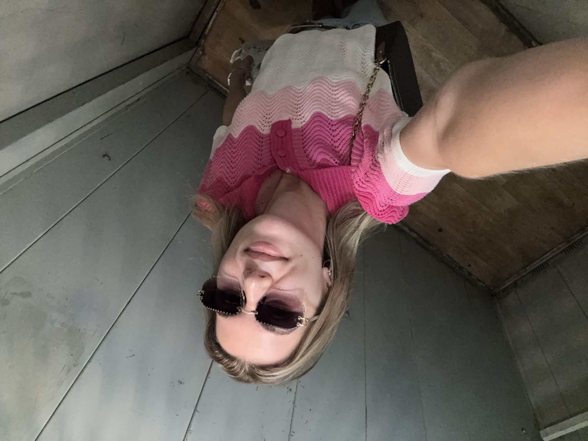 A person is taking a selfie in a narrow, enclosed space with gray, wooden paneling on the left wall and a wooden floor and ceiling. The person is wearing a pink and white zig-zag patterned cardigan and sunglasses. The person's right arm and shoulder are visible, as is their face. The image is rotated 90 degrees counter-clockwise.