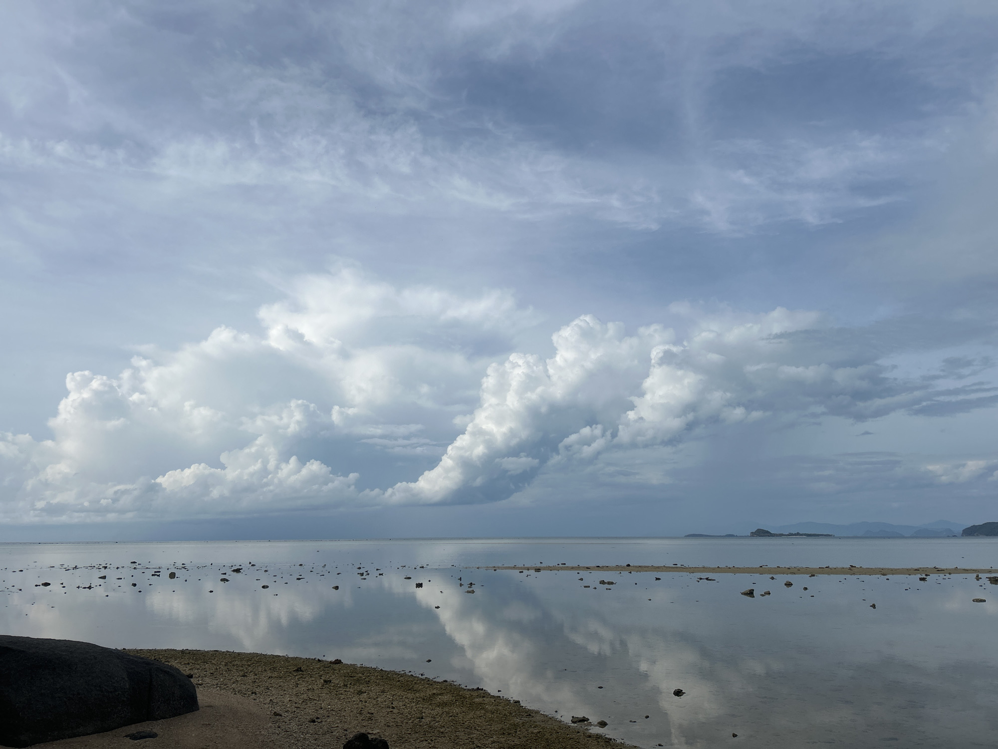 The image shows a coastal scene in Baan Harn, Thailand. The foreground features a sandy beach with scattered rocks and vegetation. A large, dark boulder is positioned at the left edge of the frame. The water is shallow and calm, reflecting the sky above. Small islands and distant mountains are visible on the horizon. The sky is dominated by heavy, gray clouds, with a large cumulonimbus formation in the center. The overall lighting suggests overcast conditions, indicating a potential impending storm.