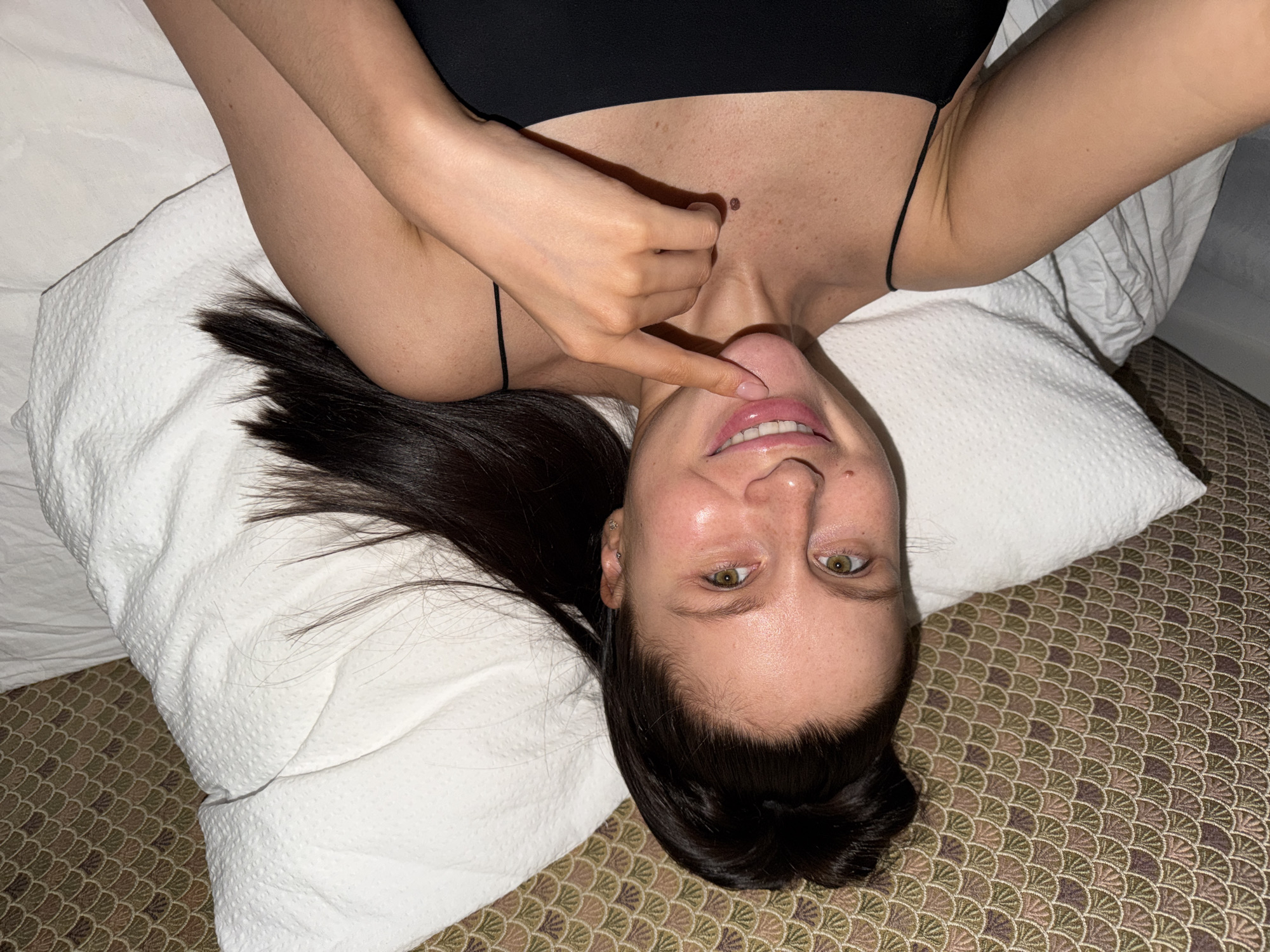 An overhead shot shows a woman lying on her back, with her head closest to the top of the frame and her body extending downwards, creating an upside-down perspective. She is looking directly at the camera with a slight smile, revealing her teeth. Her eyes are a light color, and her dark hair is splayed out around her head on a white pillow. Her face appears somewhat shiny.
She is wearing a black top or bra with thin spaghetti straps. One arm is raised, with her hand positioned near her neck and chin. Her index finger is touching the area just below her nose, and another finger is lightly touching her neck. A distinct dark mole is visible on her upper chest, above the neckline of her top.
The woman is resting on a white, textured pillow. Below the pillow, a patterned bedspread or mattress cover is visible, featuring a repeating scalloped or fan-like design in shades of brown, green, and purple. The overall scene suggests an indoor setting, likely a bedroom in Khimki, Russia. The lighting is artificial, and her expression is relaxed and playful. There is no visible text in the image.