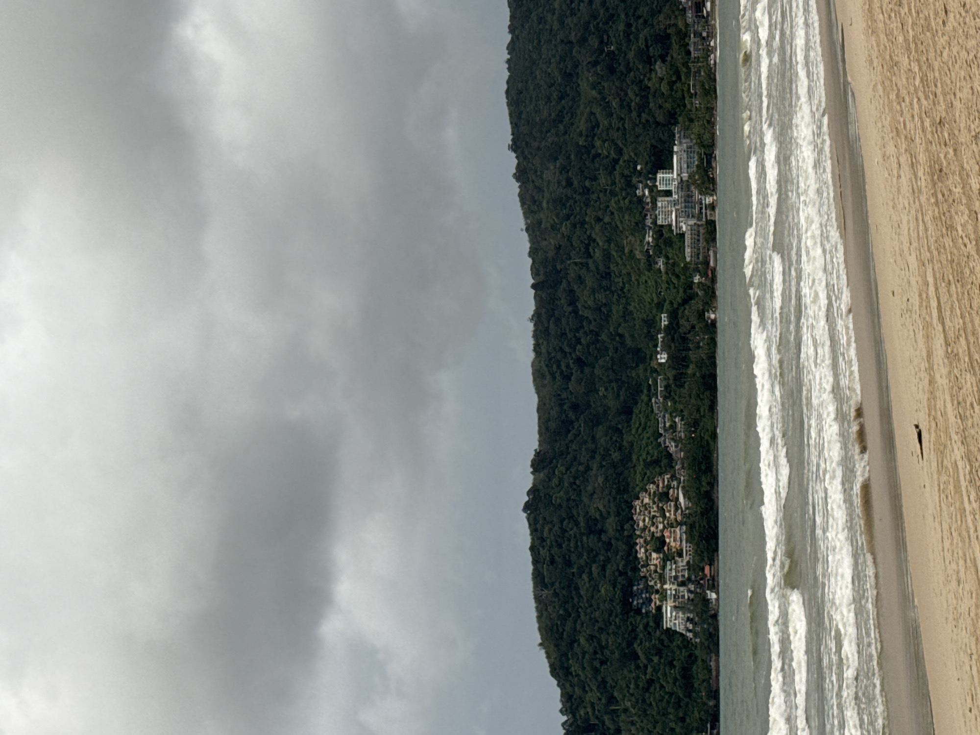 The image depicts a beach scene in Patong, Thailand, under a cloudy sky. The foreground features a sandy beach with tire tracks visible in the sand. The middle ground is dominated by the ocean, with several rows of white-capped waves rolling towards the shore. In the background, a lush green hillside rises, densely covered with trees. Along the lower slopes of the hill, a cluster of buildings can be seen, including what appears to be a resort or hotel complex with white-walled structures and a hillside village with multi-colored roofs. The sky is overcast with thick gray clouds, suggesting a potentially rainy or overcast day.