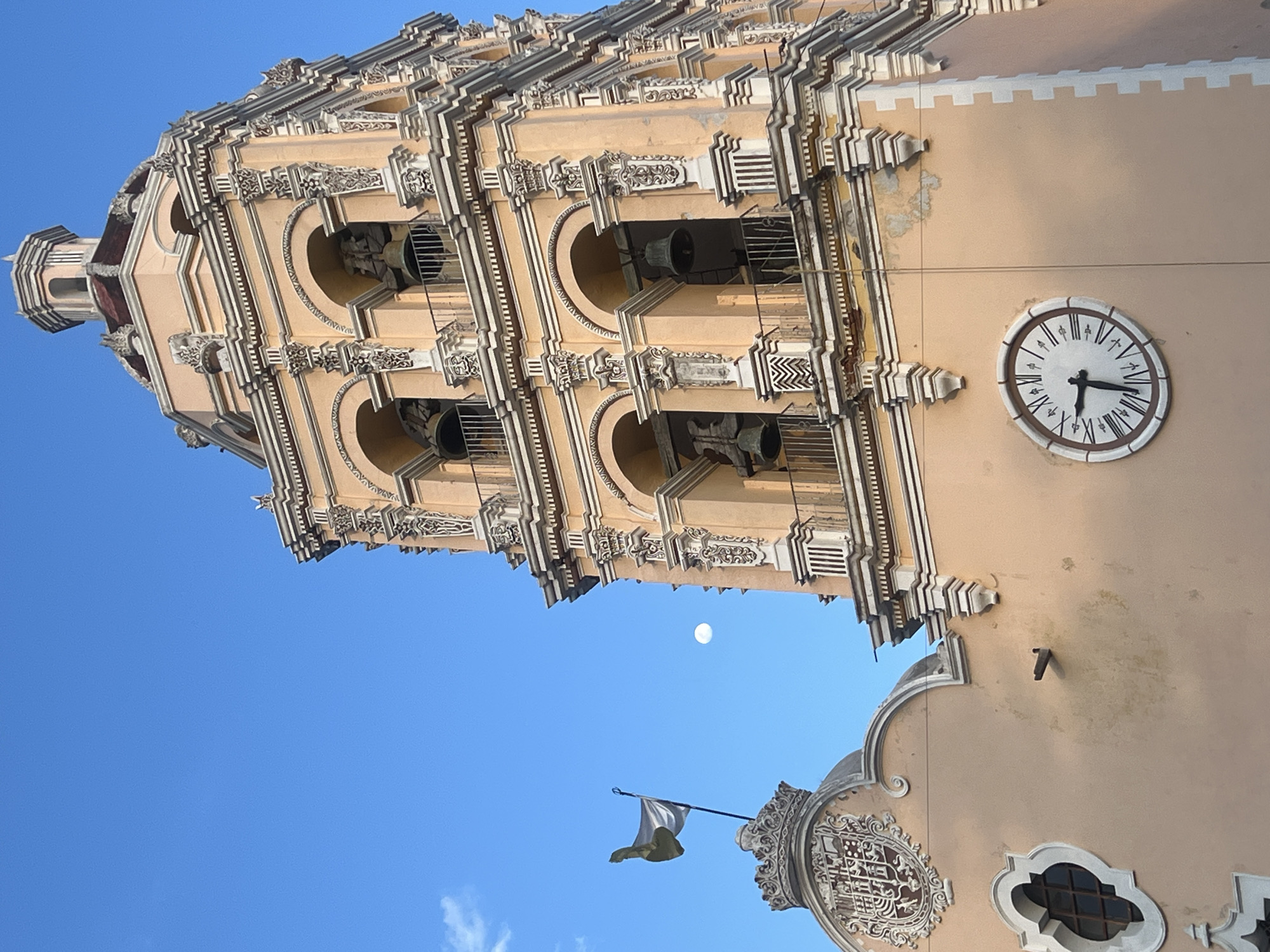 The image shows the Templo de San Francisco Acatepec, a church located in San Andrés Cholula, Puebla, Mexico. The shot is taken from a low angle, looking up at the church tower. The tower is decorated with intricate white and beige ornamentation. There are four levels of arches visible, each containing a bell. A clock is visible on the side of the tower. Below the tower, there is a decorative arched facade with a flag. A sliver of blue sky is visible in the upper portion of the image.