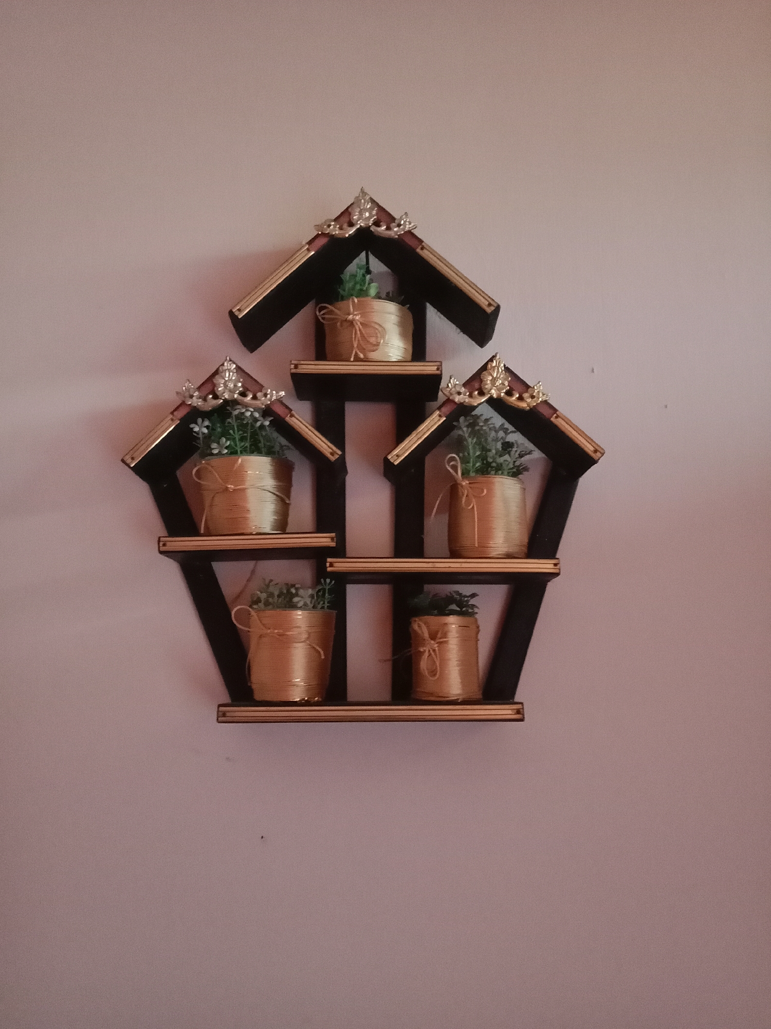 A dark-colored, multi-tiered wall shelf with a hexagonal outline is mounted on a pale pink wall. The shelf has a decorative, house-shaped top with gold trim and ornate silver accents. Each of the five shelves holds a small, gold-colored cylindrical pot. Each pot is wrapped in golden twine and tied with a simple bow. Inside each pot is a small, artificial green plant with tiny white flowers. The shelf is positioned in the center of the frame. The environment is indoor. The image does not contain any people or animals. The photograph was taken in Kutosari, Indonesia.