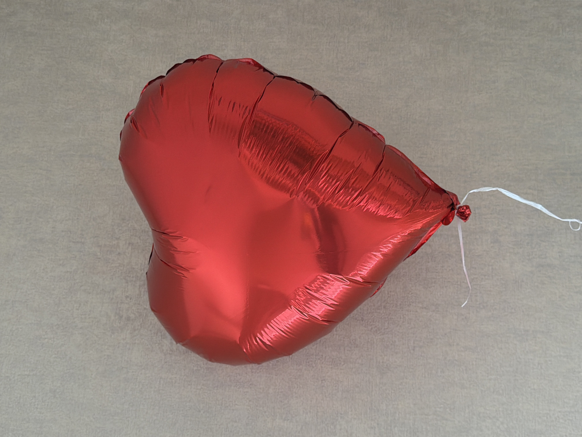 A red, heart-shaped foil balloon rests on a textured, light gray surface. The balloon is inflated and has a crinkled appearance, with a white string tied at its base. The background surface appears to be flat.