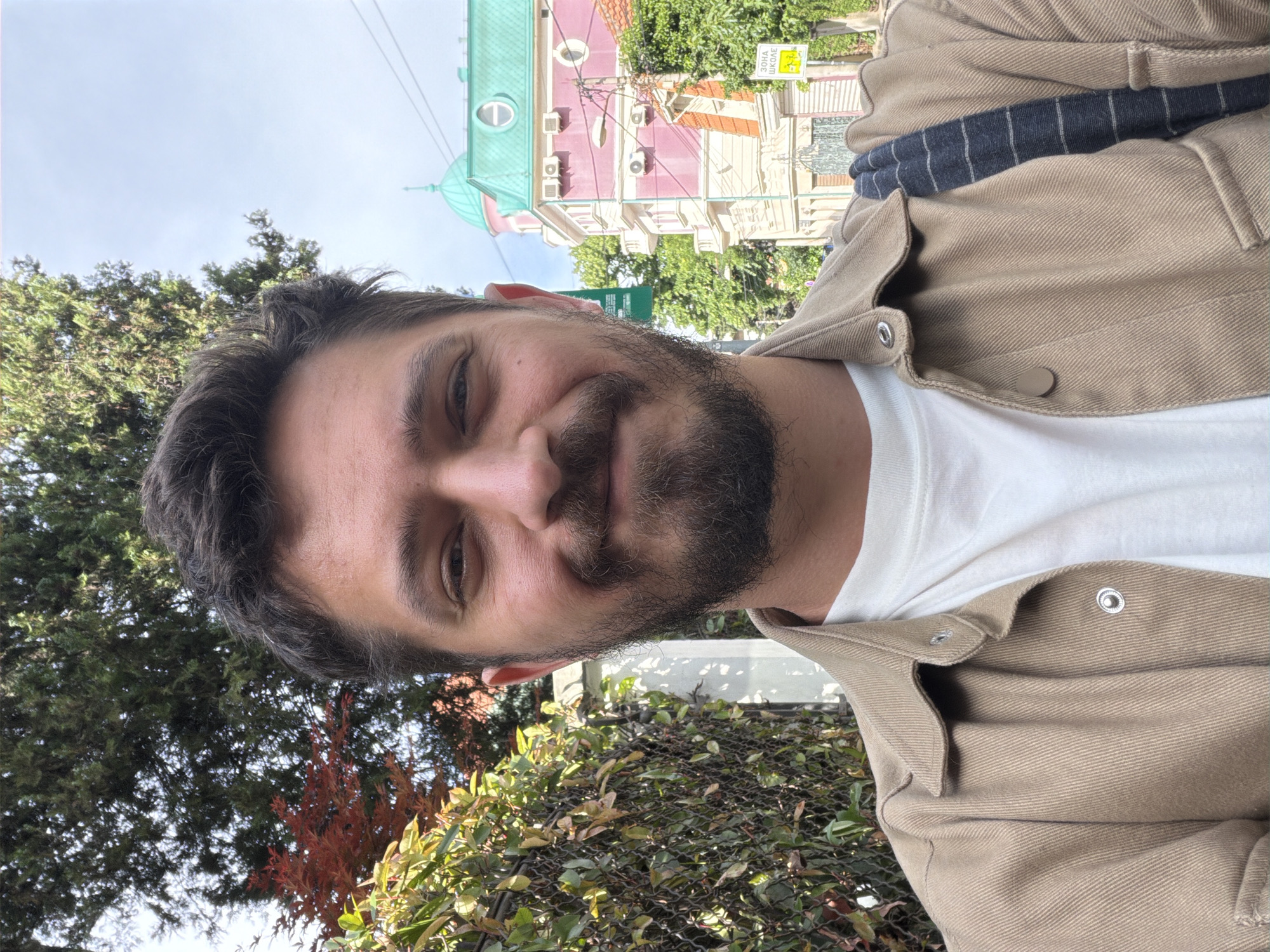 A man with a beard is seen in an eye-level selfie. He is wearing a white T-shirt and a tan jacket. Behind him are trees and foliage. In the background, there is a building with a pink facade and a green roof, along with signage in Cyrillic script, indicating a location in Belgrade, Serbia.