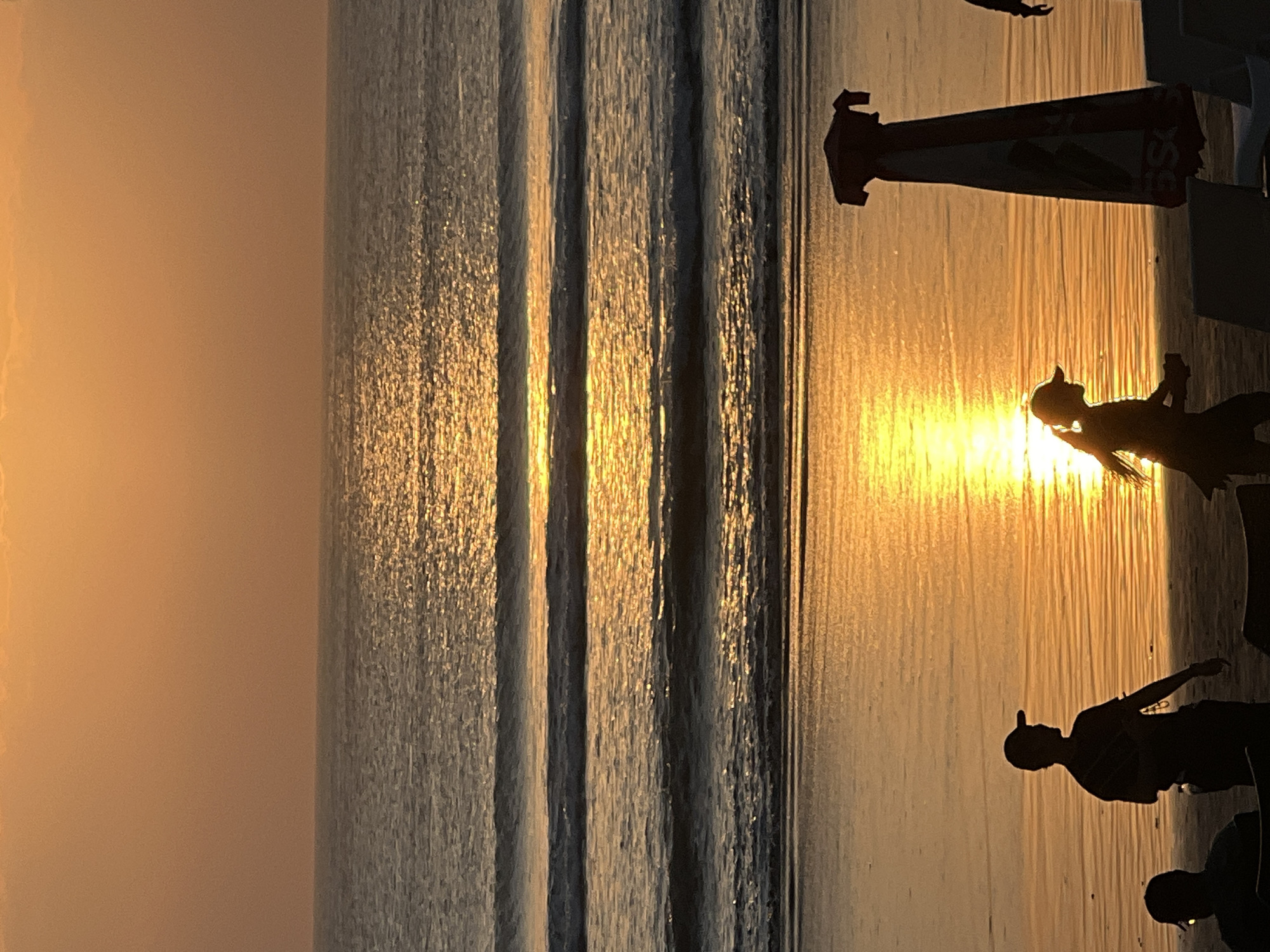 At sunset on Kuta Beach in Kuta, Indonesia, three people are visible in silhouette against the bright reflection of the sun on the ocean. A man wearing a baseball cap stands on the left, facing the ocean. To his right, a child with a ponytail and a baseball cap is walking, also facing the ocean. Further to the right, a closed beach umbrella is standing, and a portion of another person is visible on the far right edge of the frame. The ocean exhibits several distinct waves rolling towards the shore, and the wet sand reflects the warm orange and yellow hues of the setting sun. The sky is a soft gradient of orange.