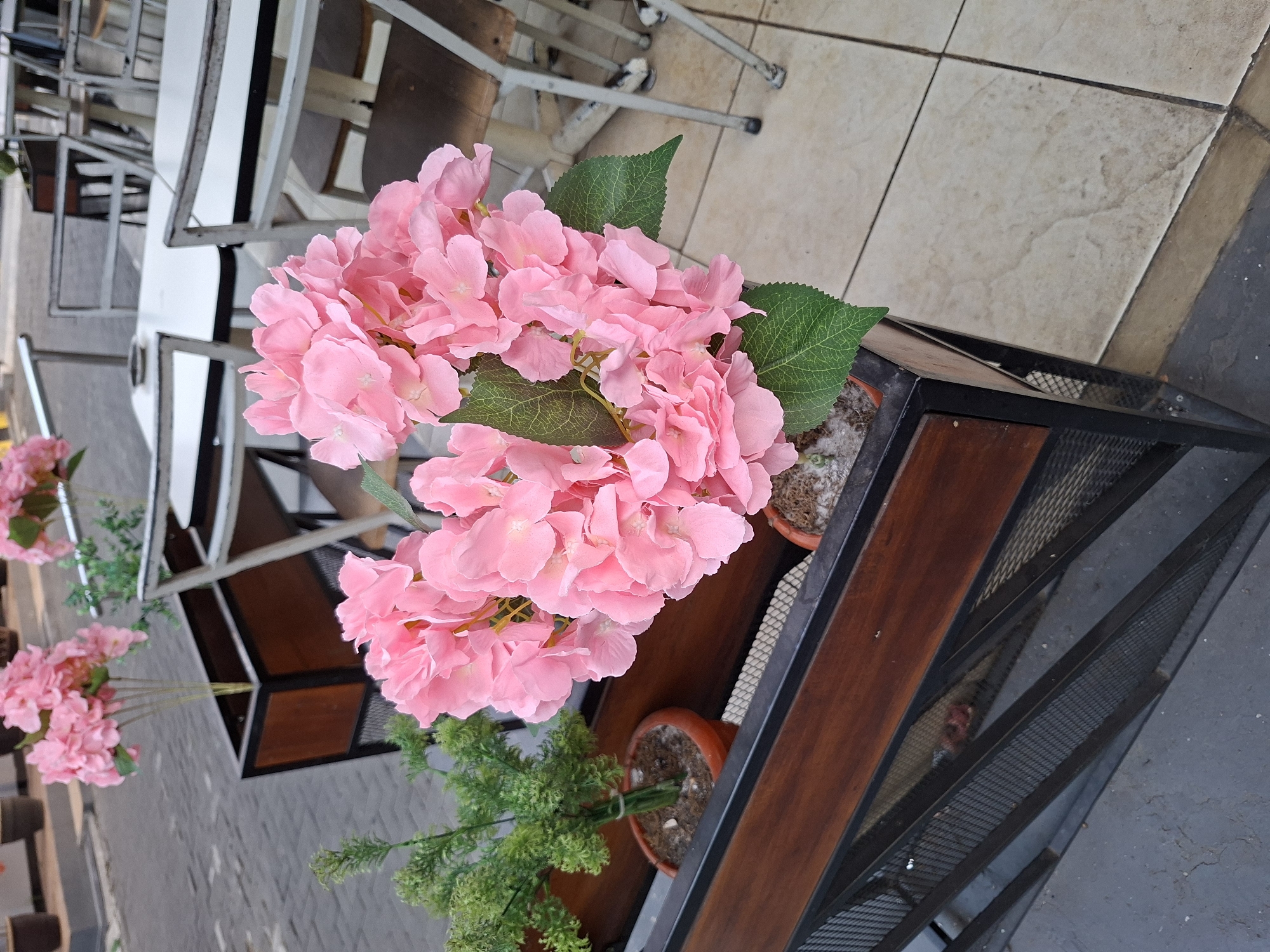 The image shows a close-up of a pink artificial hydrangea flower arrangement in a wooden planter with a metal mesh base. To the left of the hydrangea is a smaller artificial green plant in a terracotta pot. The planter is situated on a light-colored tiled surface, with a gray concrete area visible to the left and in the foreground. In the background, white tables and chairs of an outdoor cafe or restaurant are visible, along with a paved street. The lighting suggests an overcast day. The scene is likely located in a public outdoor space in Surawangi, Indonesia, possibly a street-side cafe. There are no people visible in the image.