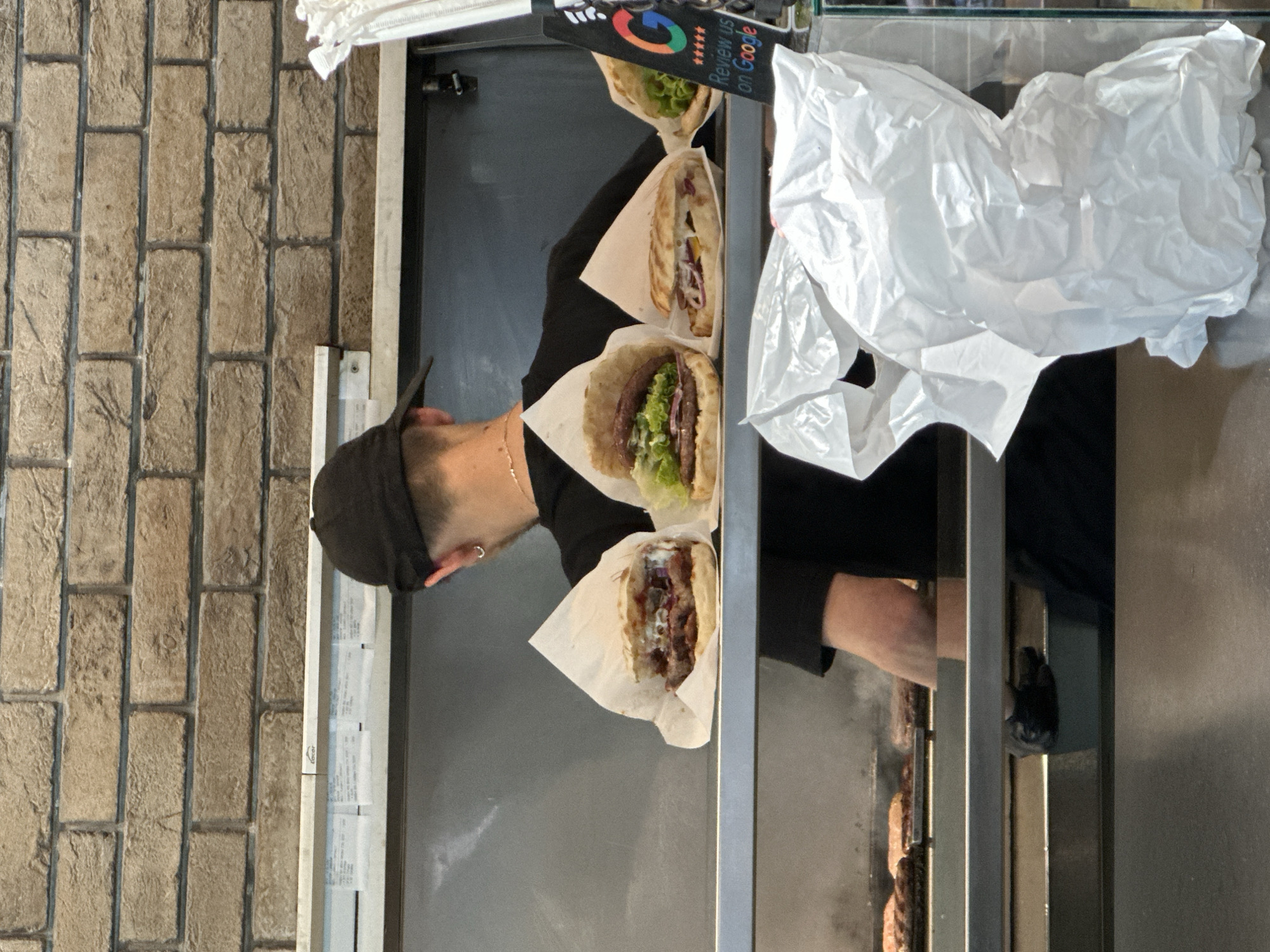 A person wearing a black cap and black long-sleeved shirt stands in a food stall. Four sandwich-like items are positioned in front of them, wrapped in white paper, appearing to be ready to serve. A white plastic bag is positioned next to the food. A sign with the Google logo and a "Review Us on Google" message is placed on top of the food. The stall has metal framing and is beside a light brown brick wall. Meat can be seen grilling vertically inside the stall. The scene is located in the city of Plovdiv, Bulgaria.