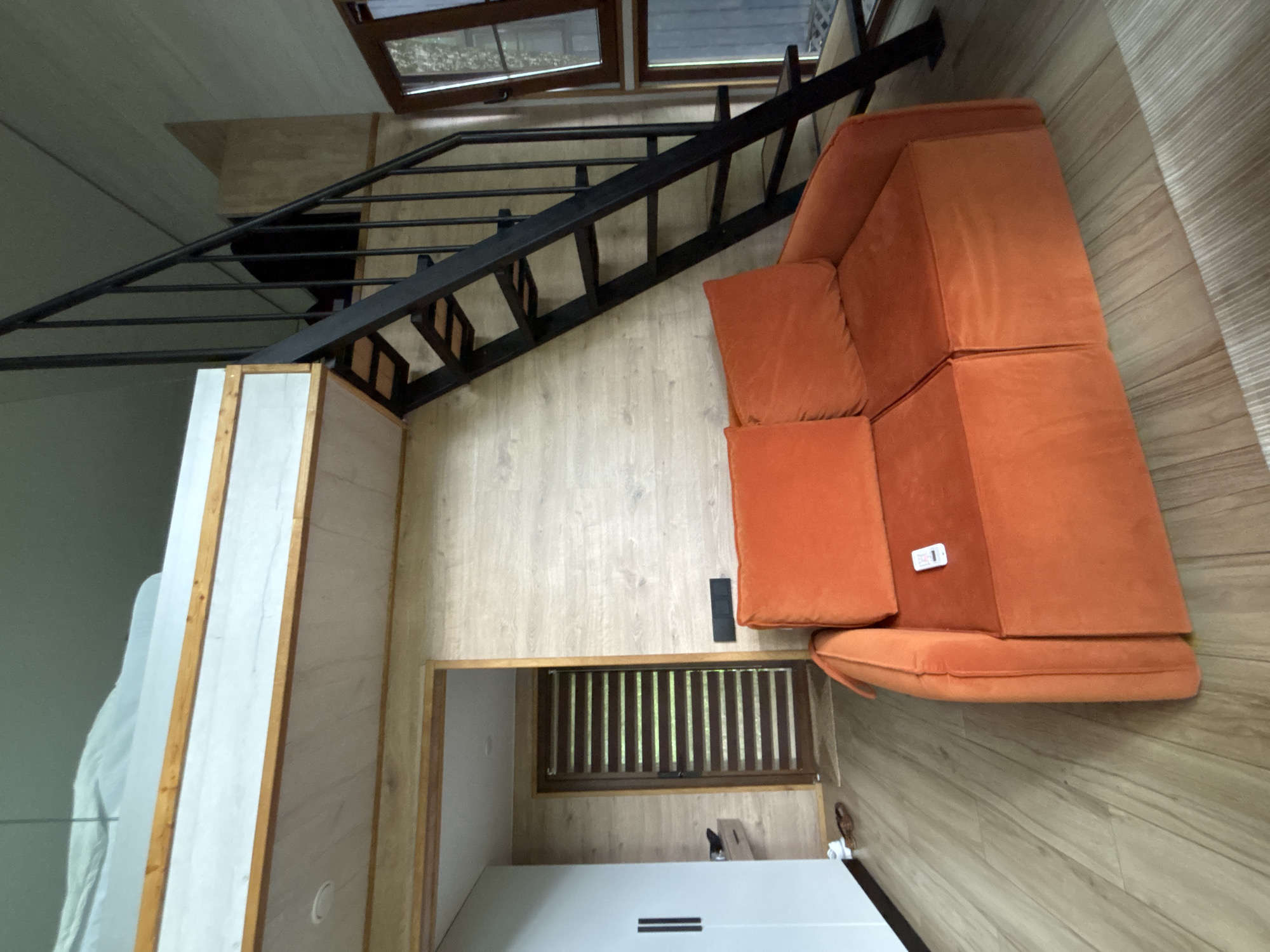 The image depicts an interior scene, likely a living space, with a prominent orange sofa in the foreground. The sofa is upholstered in a plush, possibly velvet, fabric and features two seat cushions and two back cushions. A small white remote control rests on the right seat cushion. Behind the sofa, a black metal staircase with wooden steps leads up to a lofted area, where a white mattress on a platform is visible. The walls are finished with light-colored wood paneling. To the left of the sofa, a large white wardrobe with two doors is present. A door with horizontal blinds, suggesting an exterior view of greenery, is located further back on the left. The flooring is made of light-colored wooden planks. The overall impression is a modern, compact living space with a focus on functional design. The scene is set in Krasnaya Polyana, Russia.