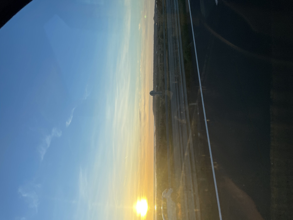 The image presents an outdoor scene at sunset, observed from a vehicle on a highway near La Palme, France. The upper two-thirds of the frame are dominated by a clear sky, transitioning from bright blue overhead to a prominent orange and yellow gradient at the horizon. The sun, a brilliant yellow orb, is positioned intensely on the far left at the horizon, casting a strong glare and reflecting brightly on the water below. A faint airplane contrail is visible in the middle-left sky.
Below the horizon, a calm body of water, likely a coastal lagoon or étang, stretches across the mid-ground, mirroring the sky's colors and the sun's brilliance. Beyond this water, a low-lying, vegetated landscape extends horizontally, characterized by scrubland and distant, slightly elevated terrain on the right. A circular road sign, featuring a white face and a dark border, stands within this vegetated area on the right side of the highway.
In the foreground, a multi-lane asphalt highway runs from left to right, parallel to the horizon. The dark asphalt surface displays white lane markings, including a solid white line defining the shoulder. A metal crash barrier, consisting of two horizontal rails, separates the highway from the adjacent landscape. A light-colored (possibly white or silver) SUV or crossover vehicle is partially visible on the far left of the highway, traveling in the same direction as the vantage point, illuminated by the setting sun. Reflections, indicative of a window, are visible across the lower portion of the image, including a dark, amorphous shape in the lower right corner. No individuals are explicitly discernible.