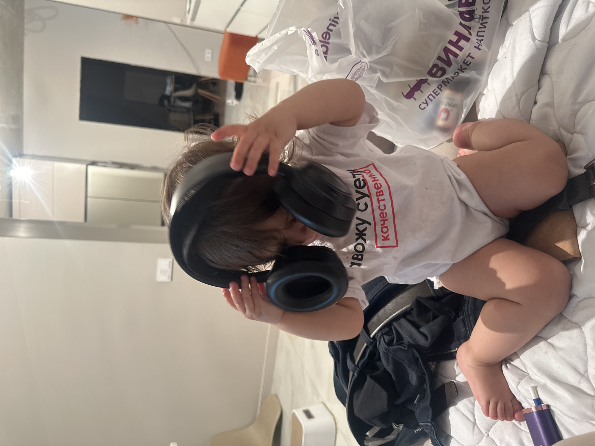 A young child, likely a baby or toddler with brown hair, is captured in a playful moment indoors. The child is positioned on a white quilted blanket, wearing a white short-sleeved bodysuit or t-shirt. They are actively engaged with a pair of large, black over-ear headphones, holding them with both hands as if attempting to put them on or exploring them.
Next to the child, a white plastic shopping bag from a supermarket is visible. Text on the bag reads "ВИНАДЕО СУПЕРМАРКЕТ НАПИТКОВ" (VINADEO SUPERMARKET NAPITKOV), which translates to "VINADEO DRINKS SUPERMARKET," suggesting a beverage store, fitting the Moscow, Russia context. Inside the bag, a metallic canister is faintly discernible. Crumpled dark clothing, possibly jeans, lies beside the child on the blanket. Near the child's feet, a purple and white electronic device, resembling a vape pen, rests on the blanket.
The child's white t-shirt bears red text which reads "ВВОЖУ СУЕТУ КАЧЕСТВО" (VVOZHU SUETU KACHESTVO), translating to roughly "I INTRODUCE HUSTLE QUALITY" or "I BRING FUSS/BUSTLE QUALITY."
The background features a reflective surface, likely a mirror or glossy furniture, showing indistinct reflections of an interior space, including what appears to be a dark screen or monitor and part of an orange chair. A bright light source, possibly an overhead light or window, creates a starburst reflection on the left side of the image. The scene suggests a casual, domestic environment during daytime, with the child exhibiting curiosity and playfulness.