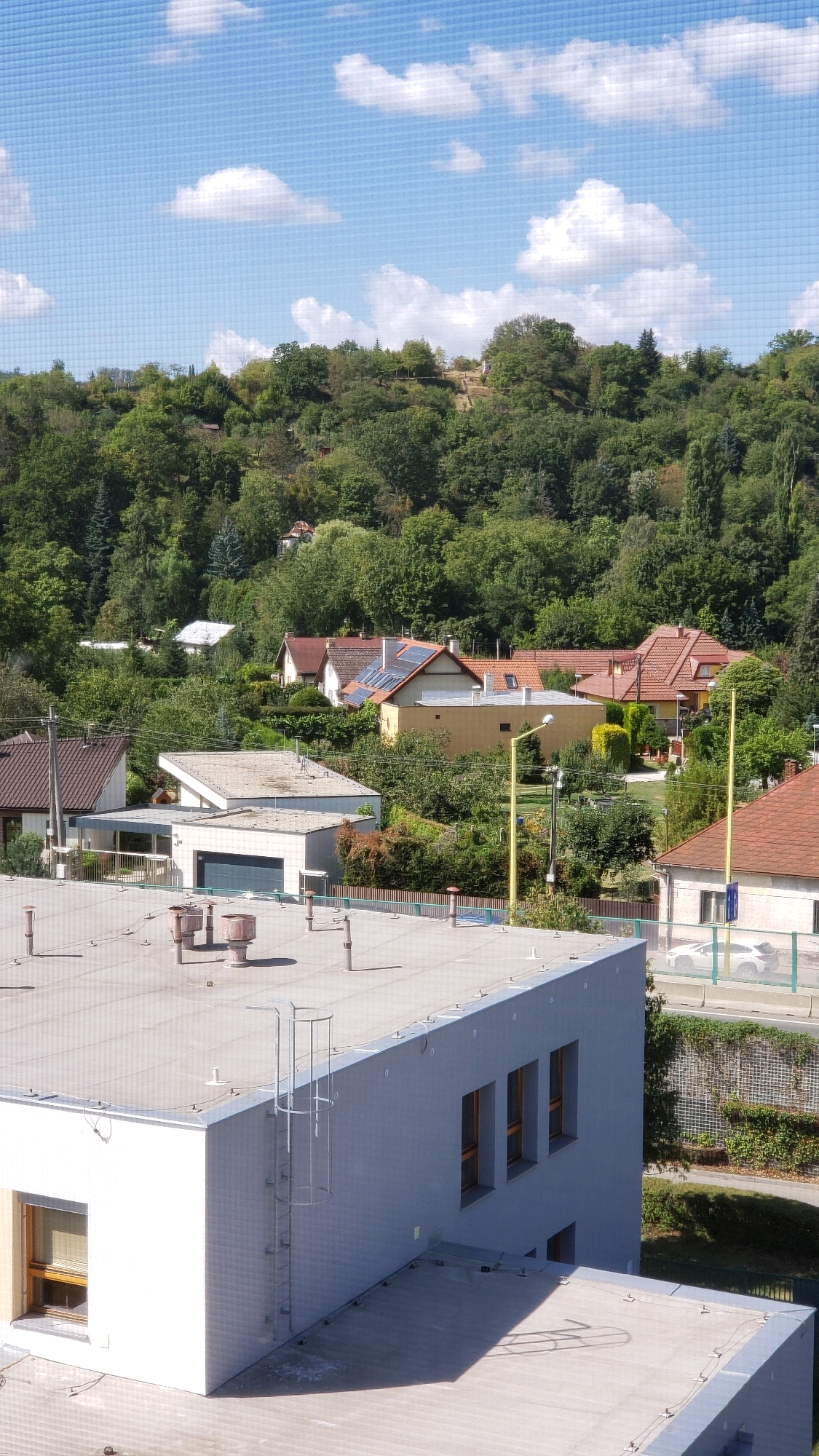 The image shows a view of a residential area with some modern buildings in the foreground and a wooded hill in the background. The sky is blue with scattered white clouds. The foreground features the flat roof of a modern, light gray building with several vents and pipes. A metal ladder extends vertically up the side of this building, which has several windows. Adjacent to this building is another structure with a white facade and a pitched roof. Further back, there are several houses with red-tiled roofs, some with solar panels. A yellow street lamp is visible in the middle ground. The background is dominated by a densely wooded hill with trees of varying shades of green. The overall scene depicts a sunny day in a suburban or semi-rural environment, likely in the city of Prešov, Slovakia, given the context. No people are visible in the image.