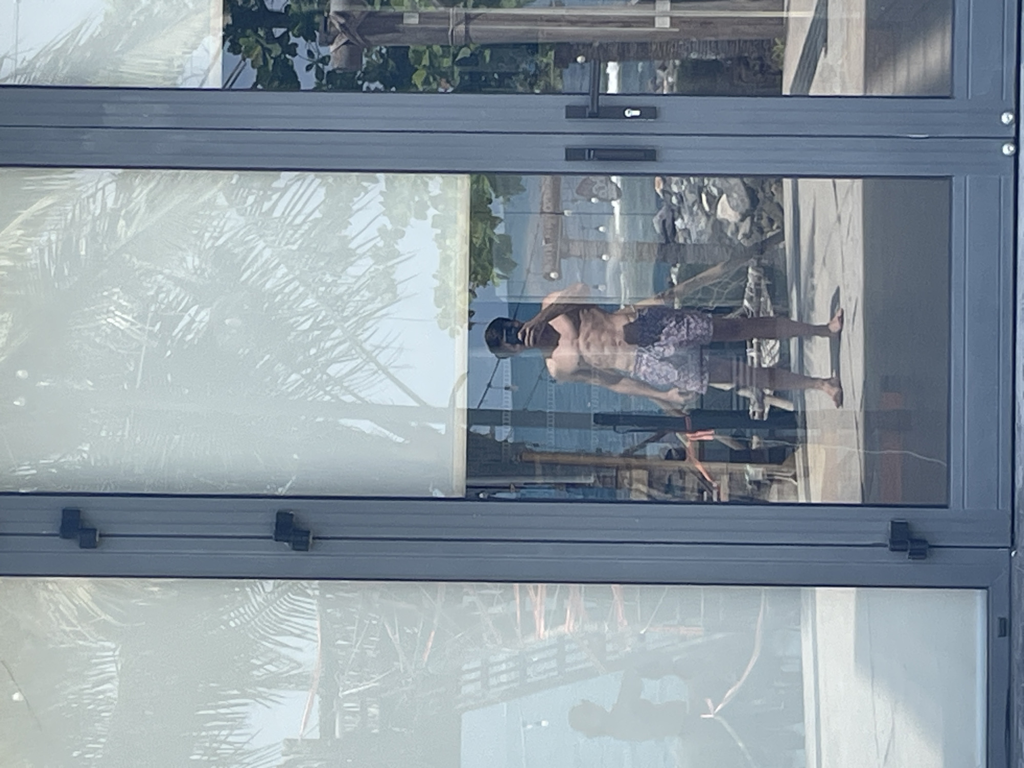 The image shows a man in swimwear reflected in a window, possibly from inside a building. The reflection also includes glimpses of the surrounding environment, suggesting a coastal or waterfront location. Other reflections are visible, including trees, buildings, and other objects. The location is identified as Bac Nha Trang Ward, Vietnam.