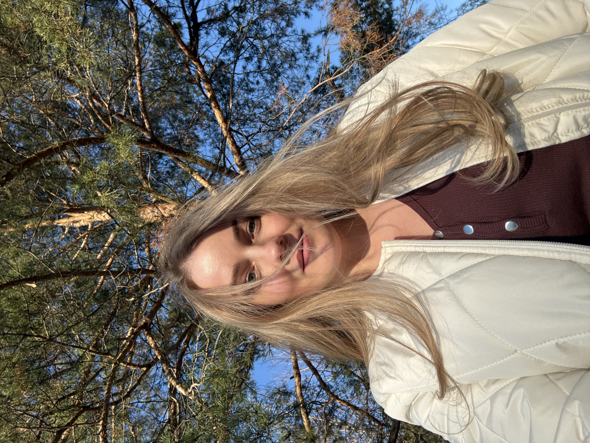 The image shows a woman with long blonde hair, wearing a burgundy top with silver buttons and a white quilted jacket. She is outdoors, seemingly in a wooded area with pine trees visible behind her. The sky is blue and there is sunlight. The location is Chaikovsky, Russia.