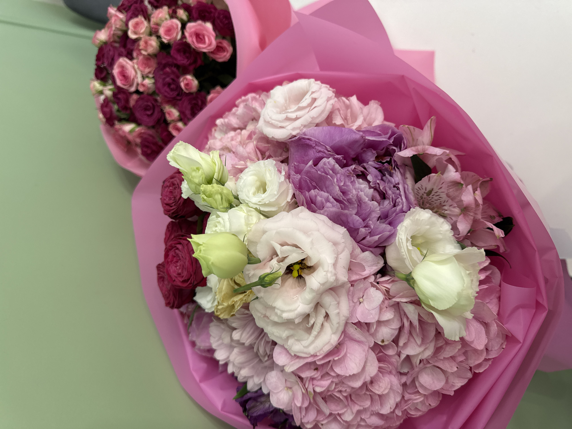 Two bouquets of flowers are displayed against a light green wall and a white surface. The bouquet in the foreground is a vibrant arrangement featuring large pink hydrangeas, a large purple peony, white eustomas, and a few pink alstroemerias. The flowers are wrapped in bright pink paper. In the background, a second bouquet is visible, composed of numerous small pink and deep magenta roses, also wrapped in pink paper. The scene appears to be indoors, possibly in a shop or a home, with no specific city or location details discernible from the image.